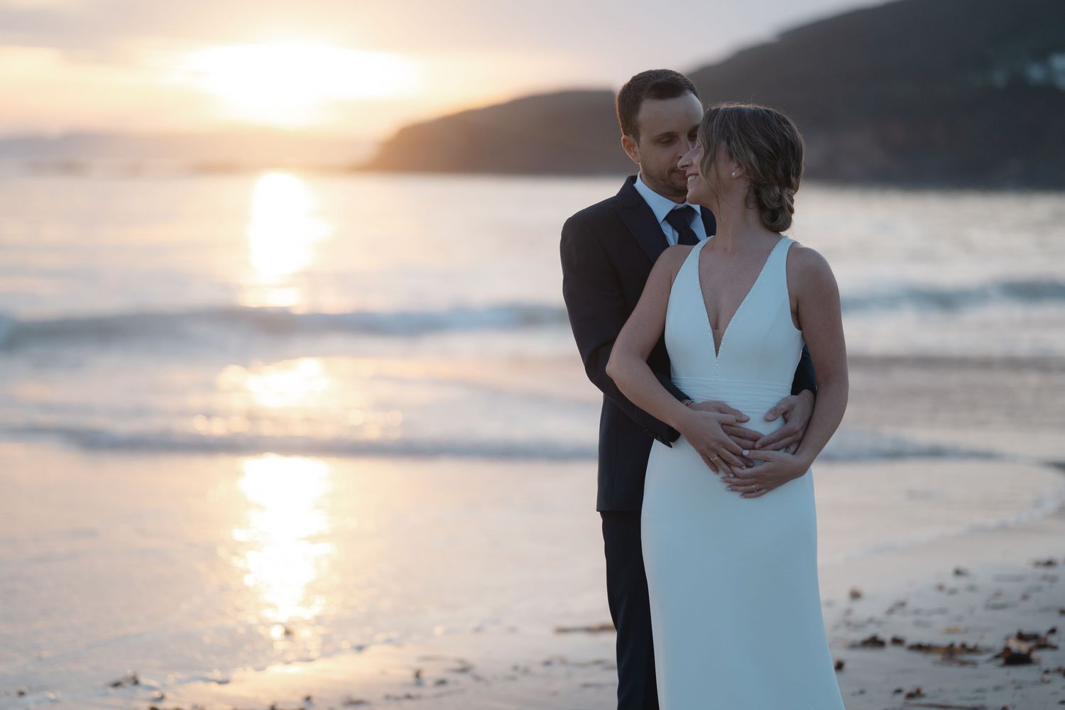 A couple shares an intimate moment on a beach at sunset with golden light reflecting on the ocean.