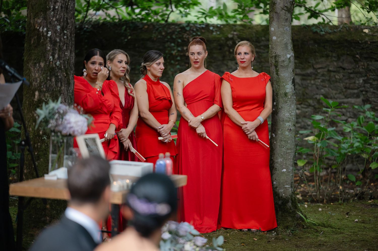 A group of bridesmaids wearing matching red dresses stand together at an outdoor wedding ceremony.