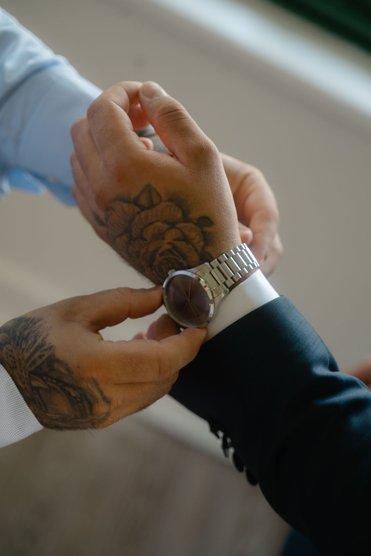 Tattooed hands adjusting a watch on a wrist while wearing formal attire.
