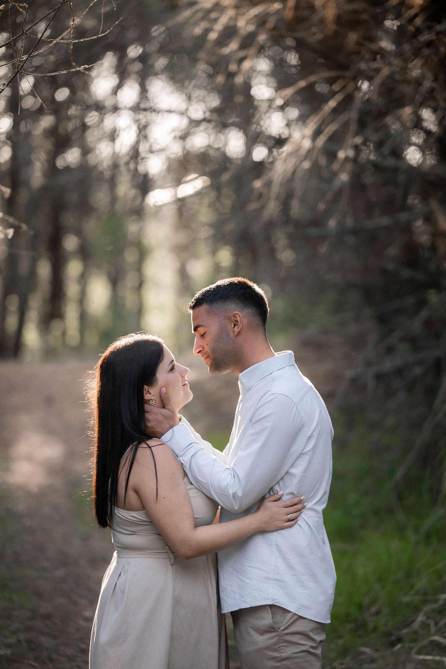 A romantic couple embraces on a sunlit forest path with dreamy bokeh lighting in the background.