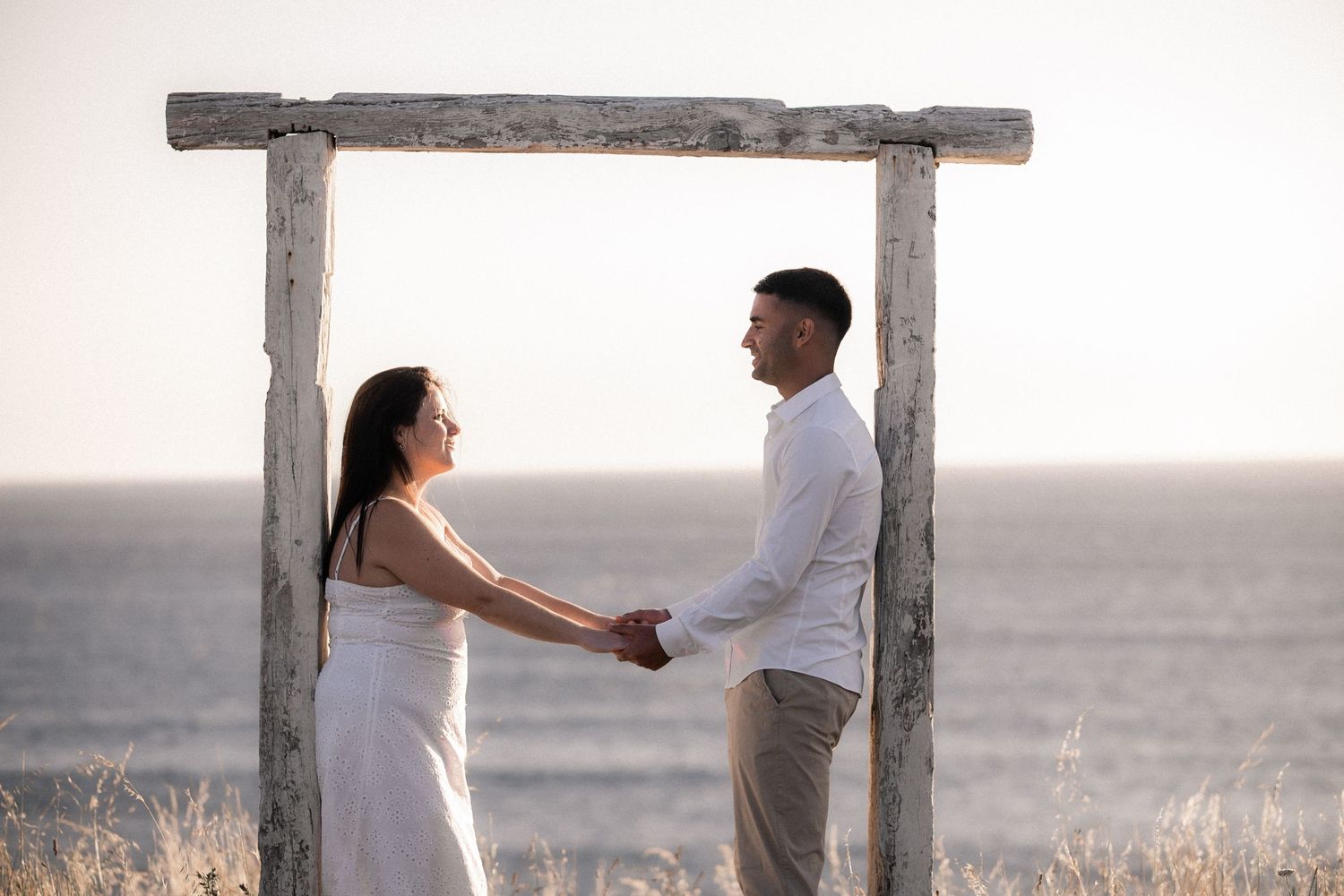 A couple shares a romantic moment under a wooden arch during sunset at an outdoor location.