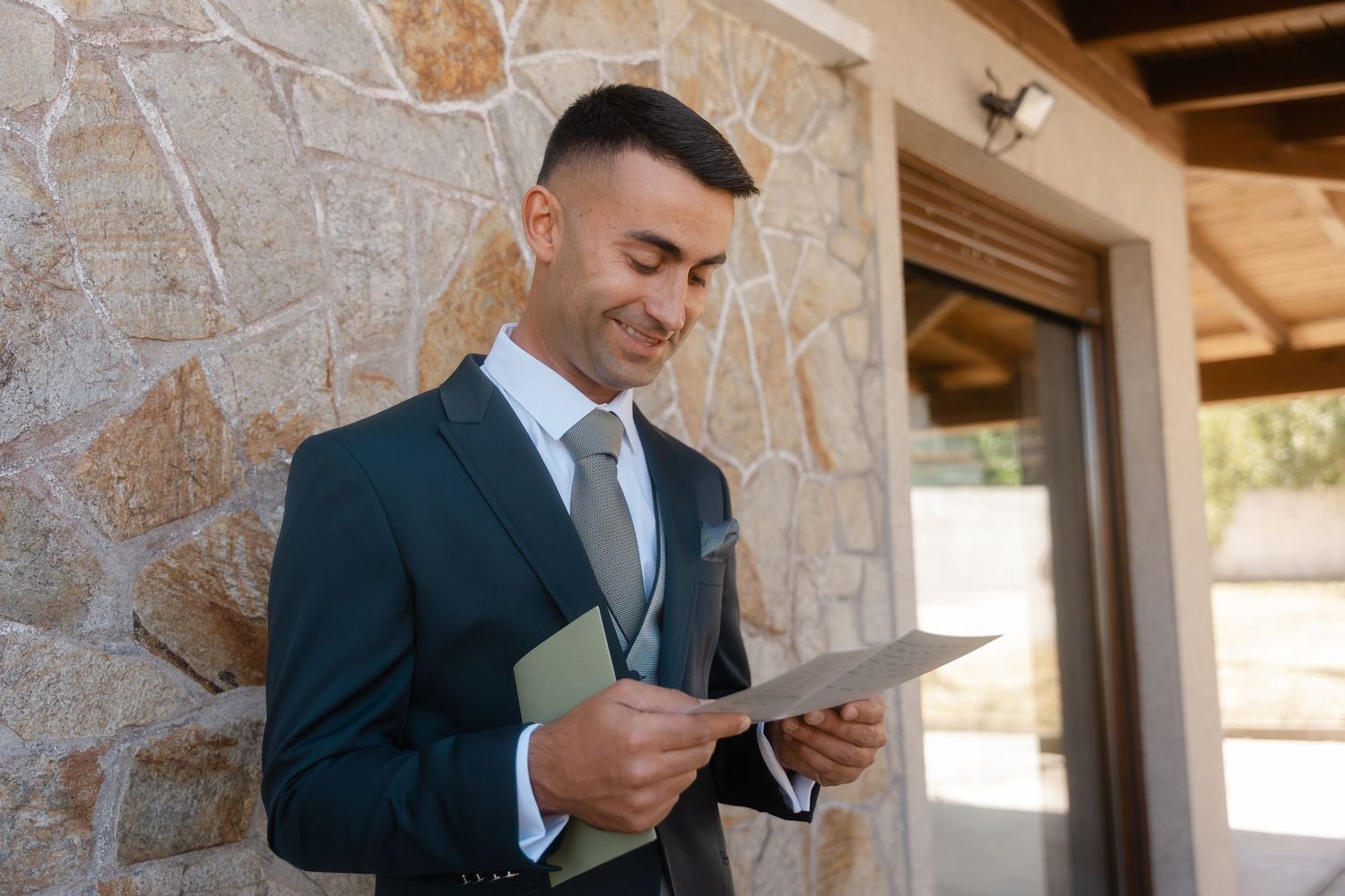 Man in dark suit looking at document while standing near stone wall in outdoor setting.