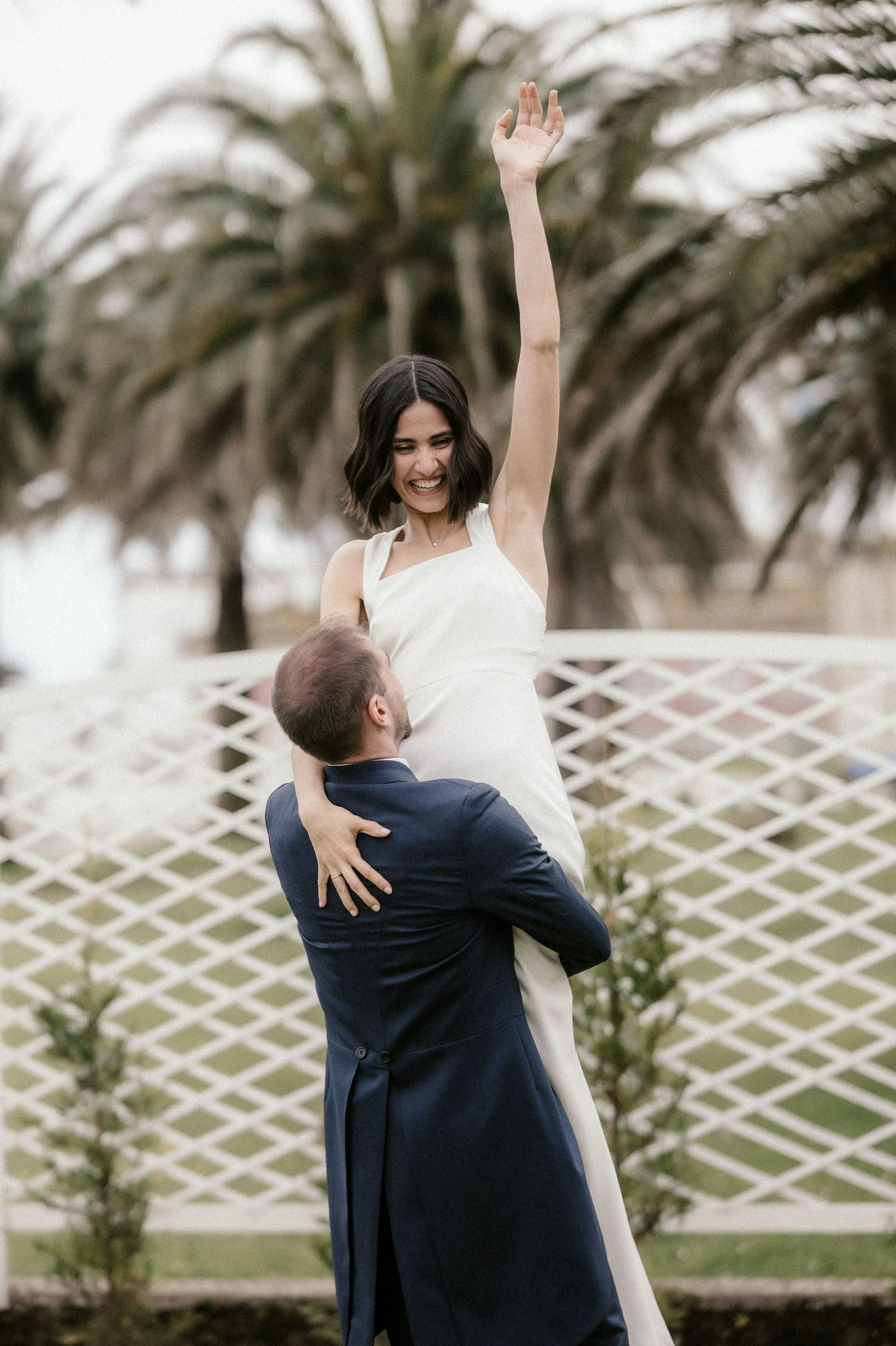 A joyful wedding moment captured as a couple embraces by a palm tree lined fence.