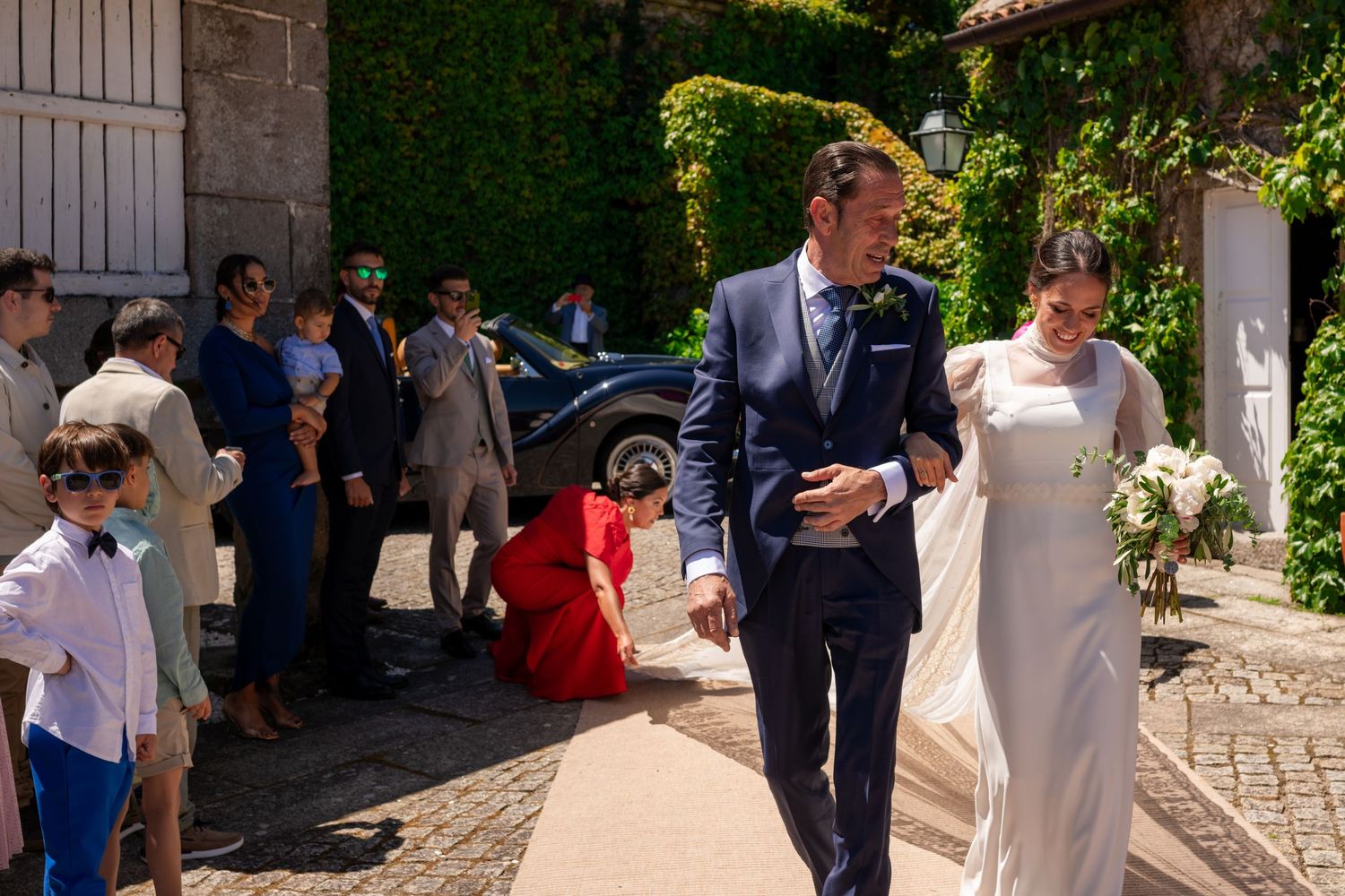 Wedding guests gather outside a rustic stone building during a sunny outdoor celebration.