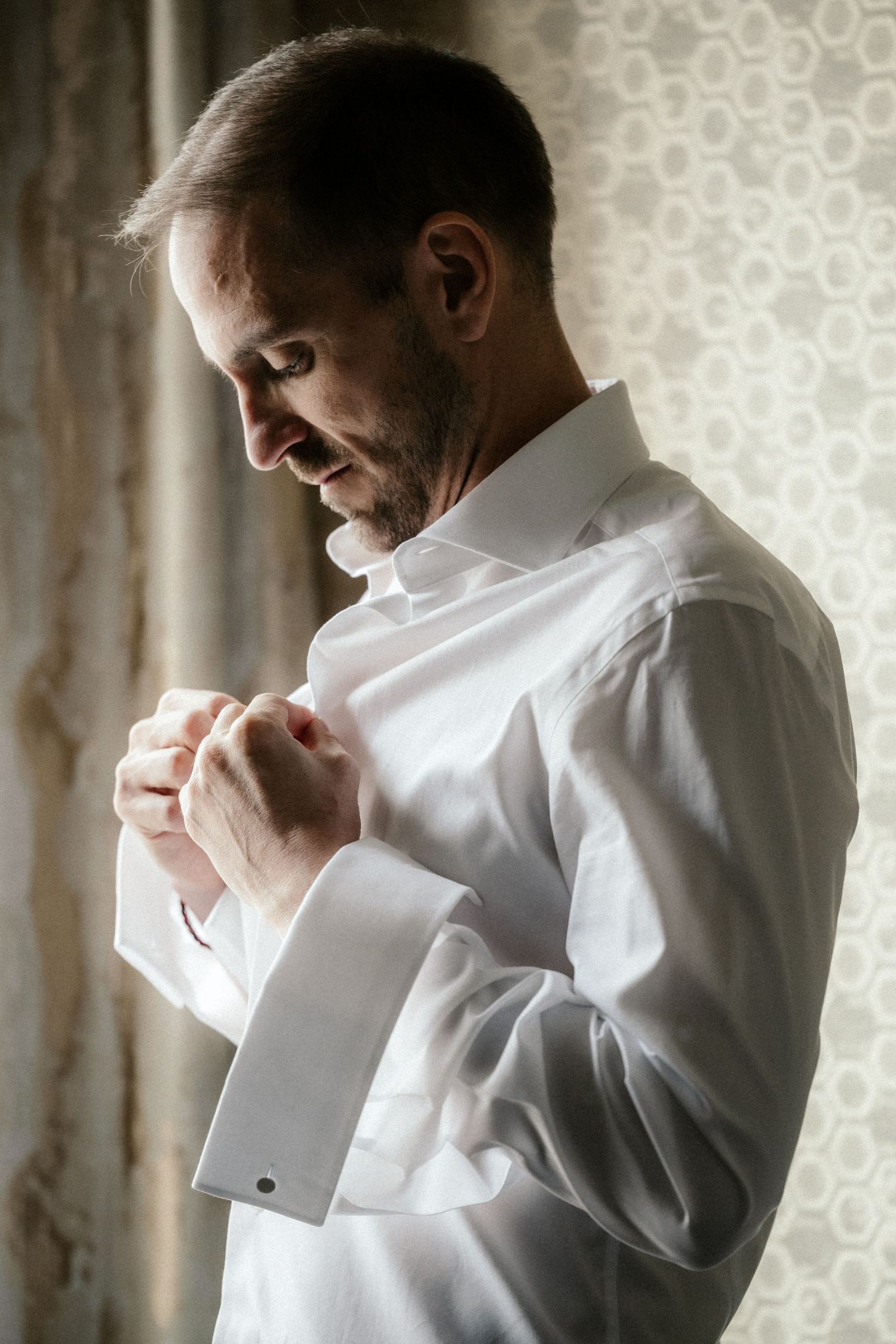 Man in white dress shirt adjusts his cuff links while getting ready against a patterned wallpaper background.