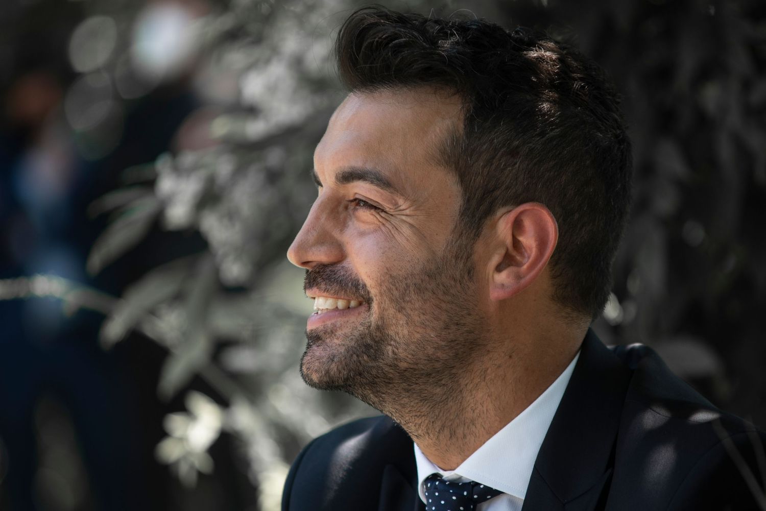 A smiling person in a dark suit and tie photographed in natural lighting with a blurred background.