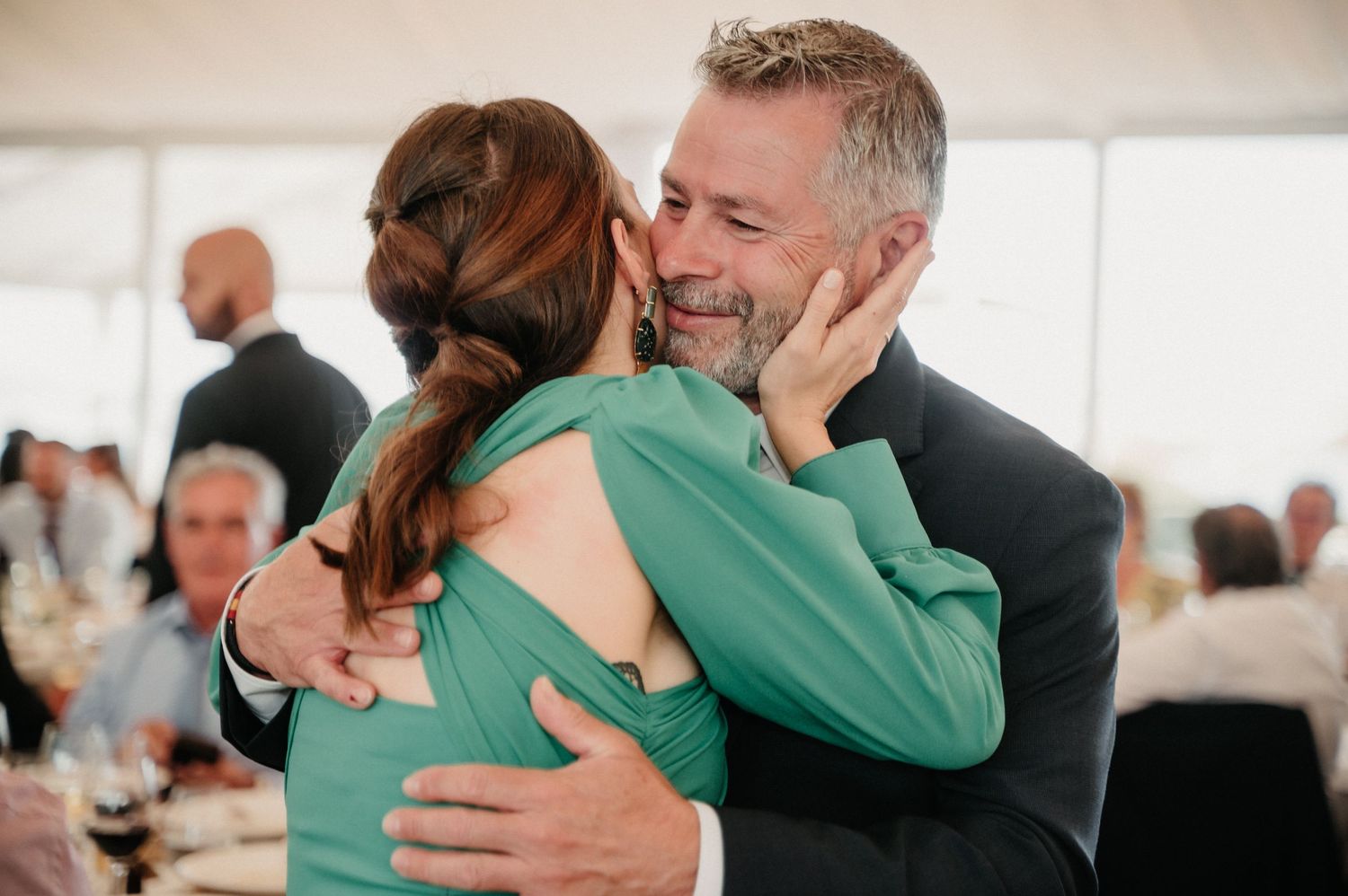 People in formal attire share warm hugs at an indoor event with natural lighting.