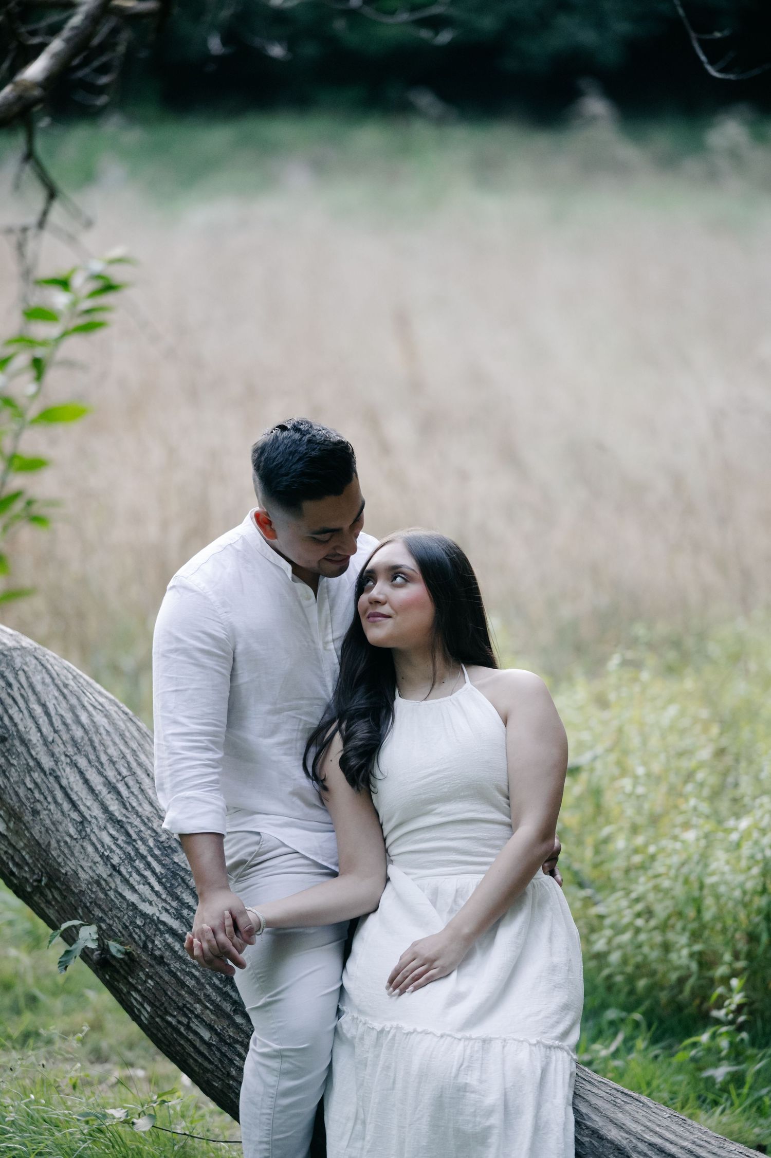 A couple in white outfits share an intimate moment while sitting on a fallen tree in a meadow.