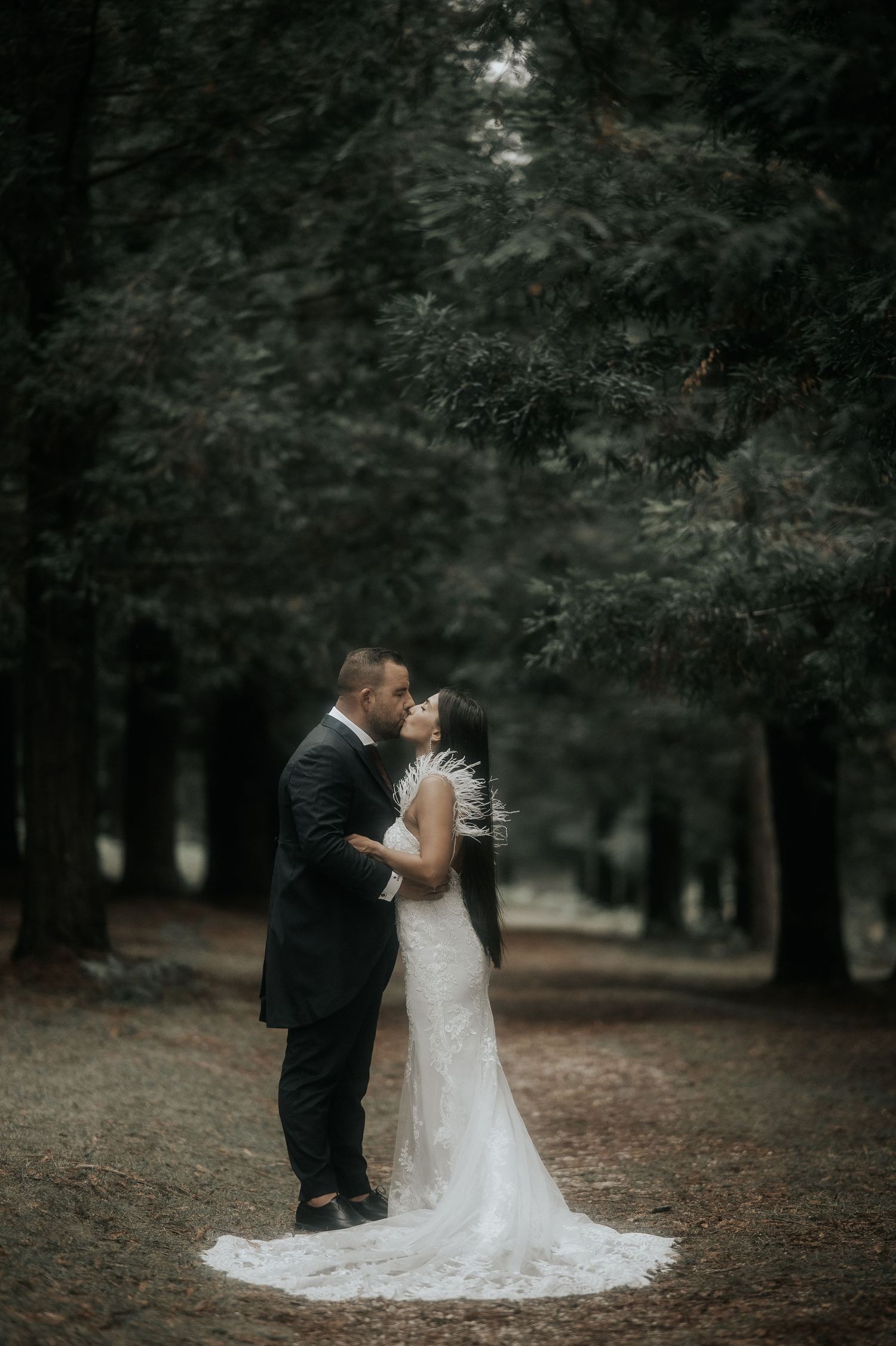 A romantic wedding couple embraces in a moody forest setting with the bride's white lace dress trailing behind.