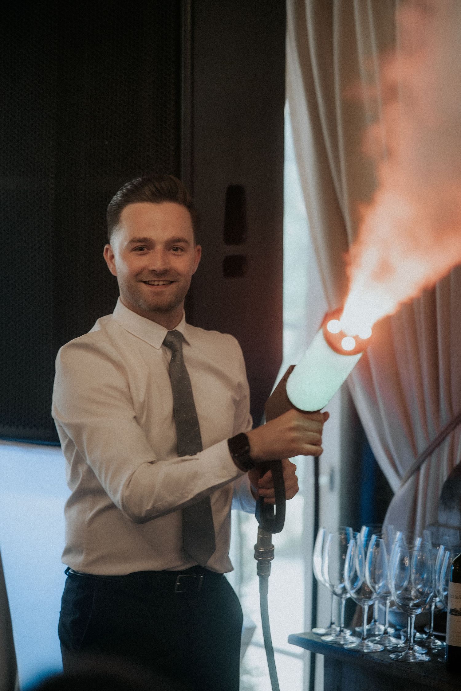 Professional in formal attire operates a fog machine next to wine glasses at an elegant event setup.