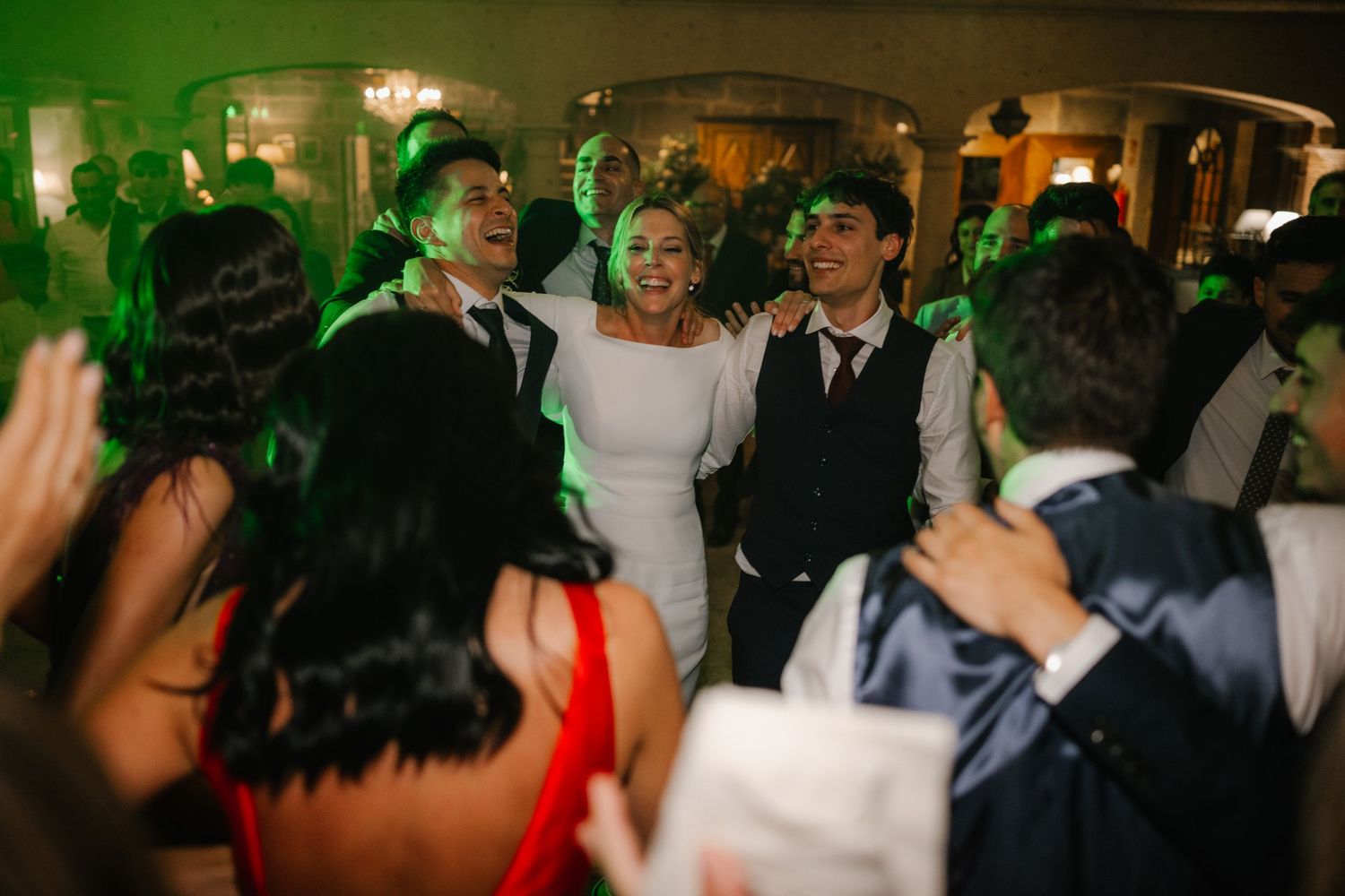 Wedding party dances energetically under moody green lighting at an indoor reception venue.