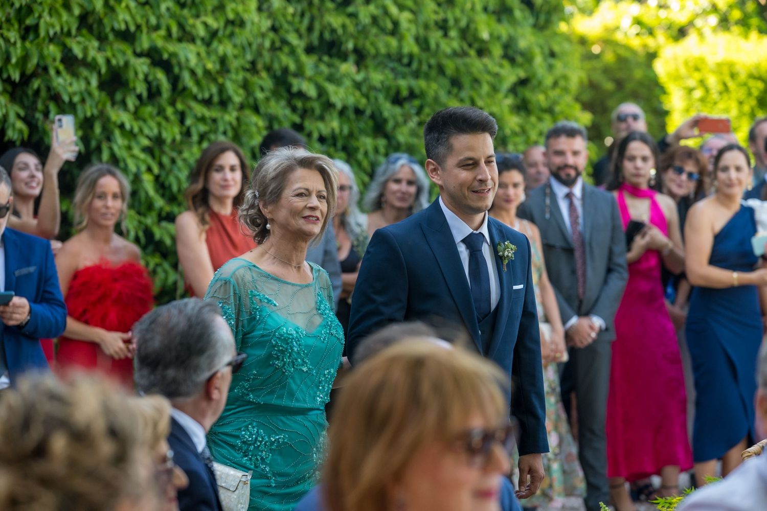 A group of wedding guests in formal attire gather outdoors in a garden setting during a summer celebration.