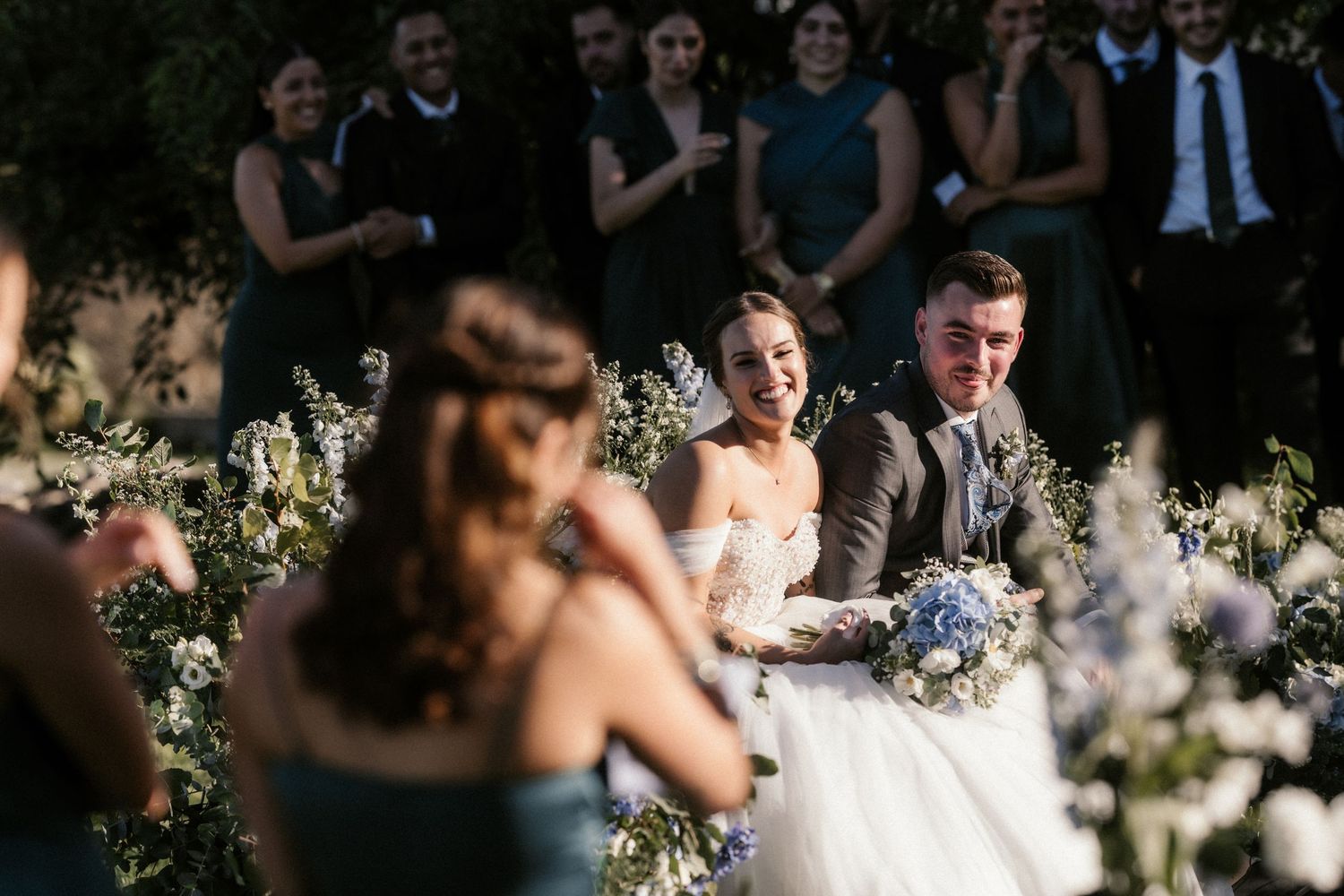 Wedding guests smile and laugh during an outdoor garden reception surrounded by white floral decorations.