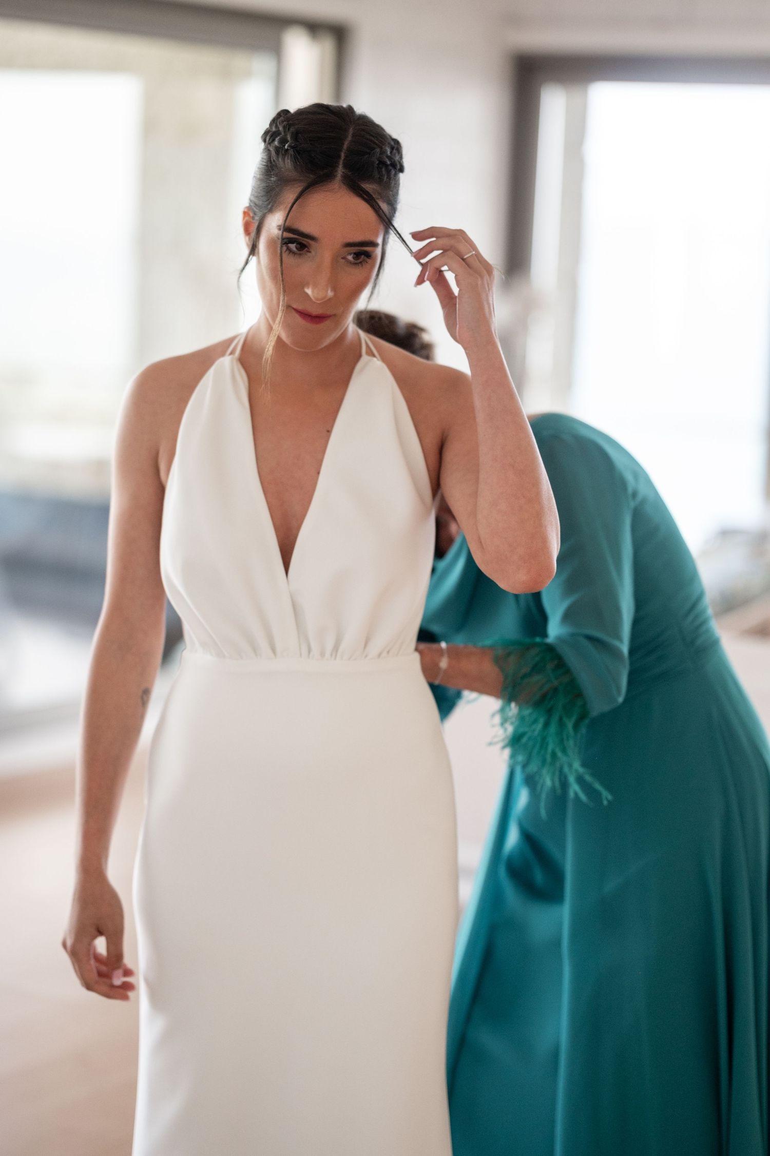 A bride in an elegant white halter wedding dress gets fitted while someone in teal assists with final adjustments.