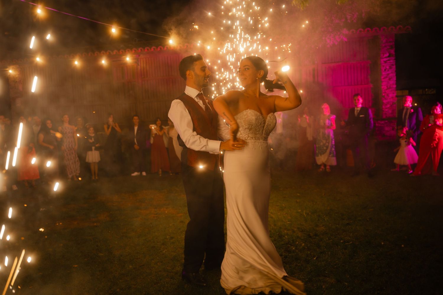 Sparklers illuminate a nighttime outdoor celebration as guests gather under string lights.