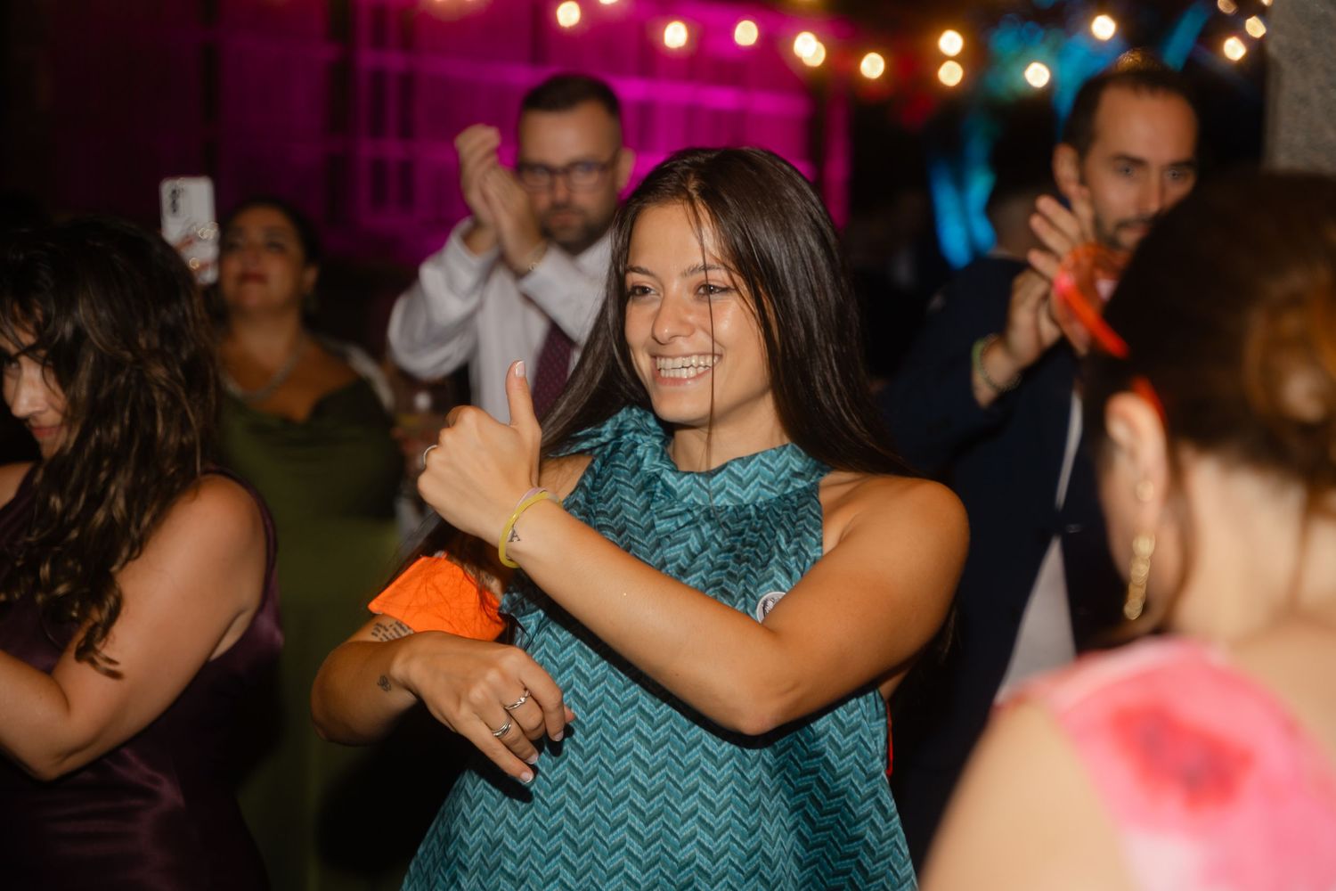 A guest in a teal chevron dress dances enthusiastically at a party with festive lights in the background.
