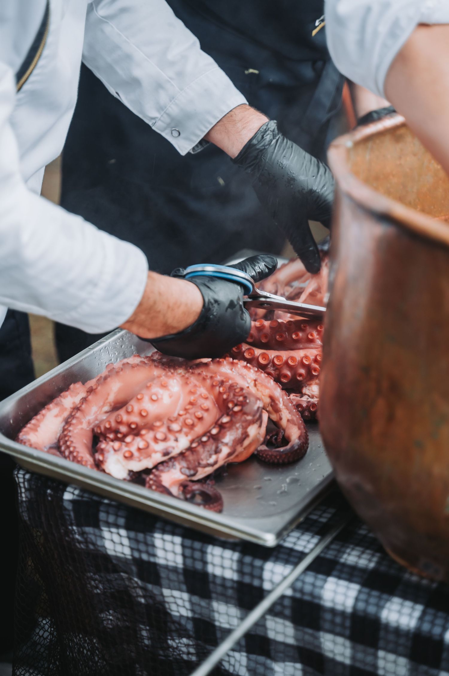 Chef in black gloves slicing fresh octopus on a checkered tablecloth at a catering station.