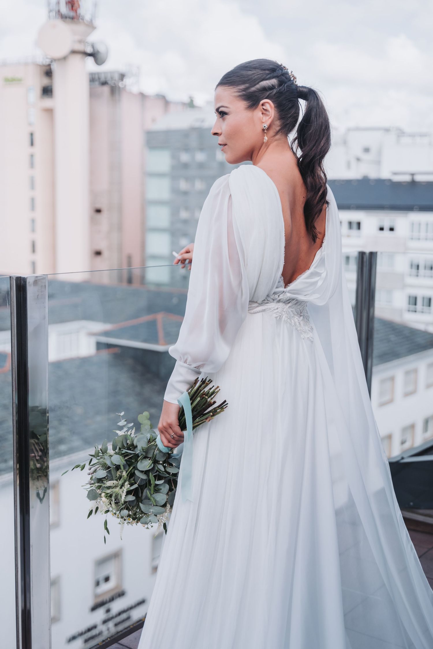 Bride in flowing white wedding dress and veil stands on balcony overlooking city skyline on cloudy day.