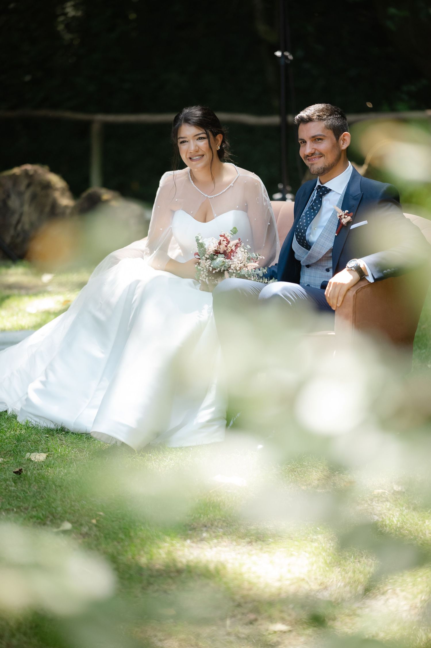 Couple sits together on grass during outdoor wedding photoshoot with soft bokeh effects in foreground.