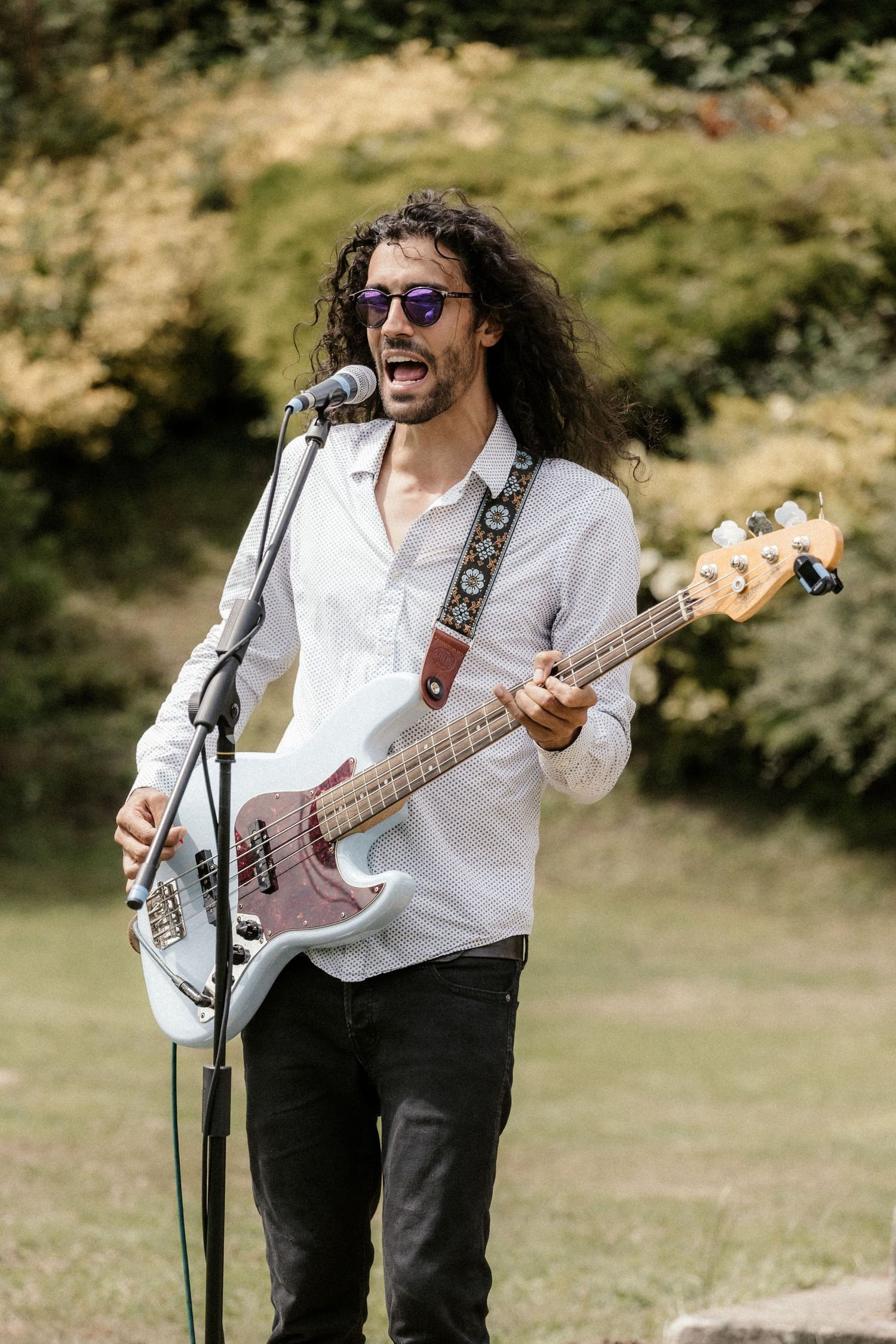 A musician in a white shirt plays bass guitar outdoors while performing at an outdoor event.