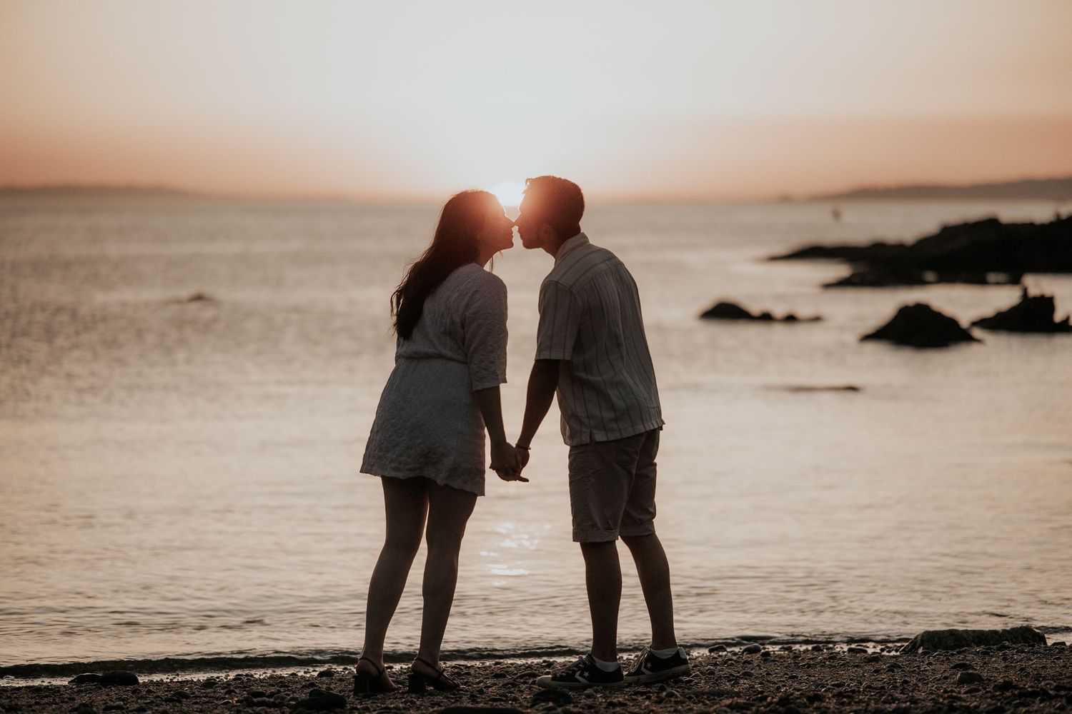 A couple holds hands while walking along a misty beach shoreline at sunset, creating a romantic silhouette.