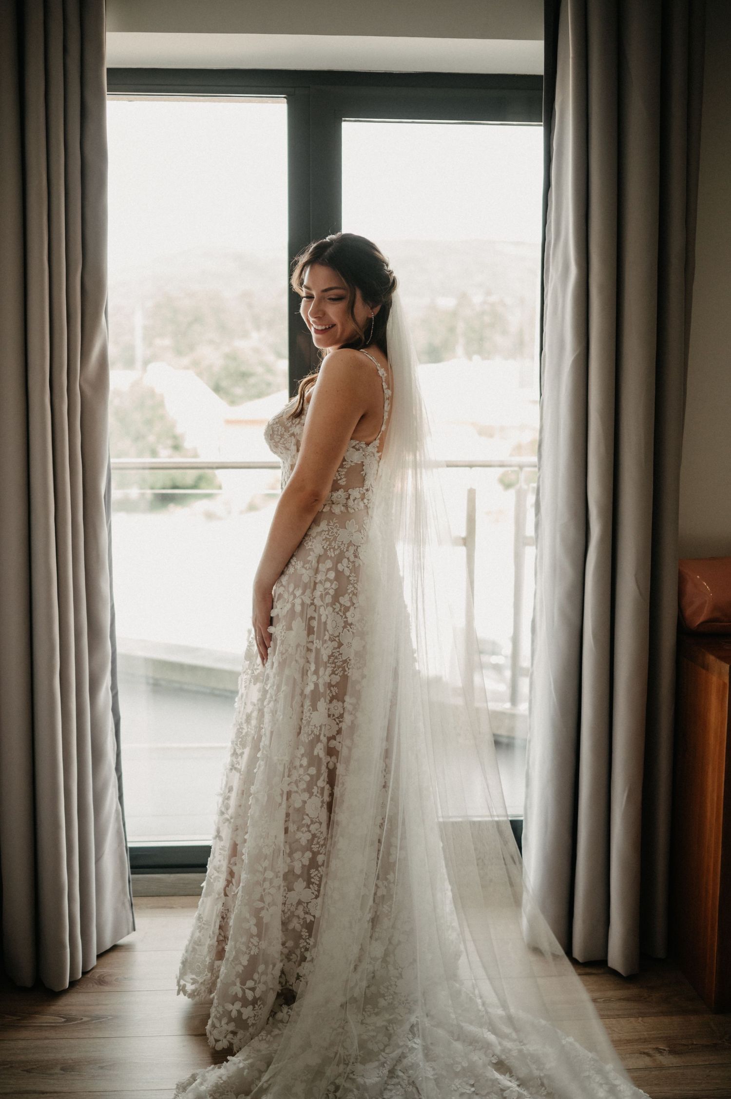 A stunning lace wedding dress with a flowing train is showcased against floor-length curtains in a bright room.
