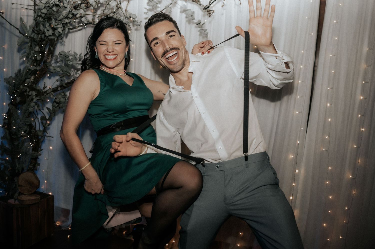 A group of wedding guests dance and pose together during a reception celebration with twinkle lights in the background.
