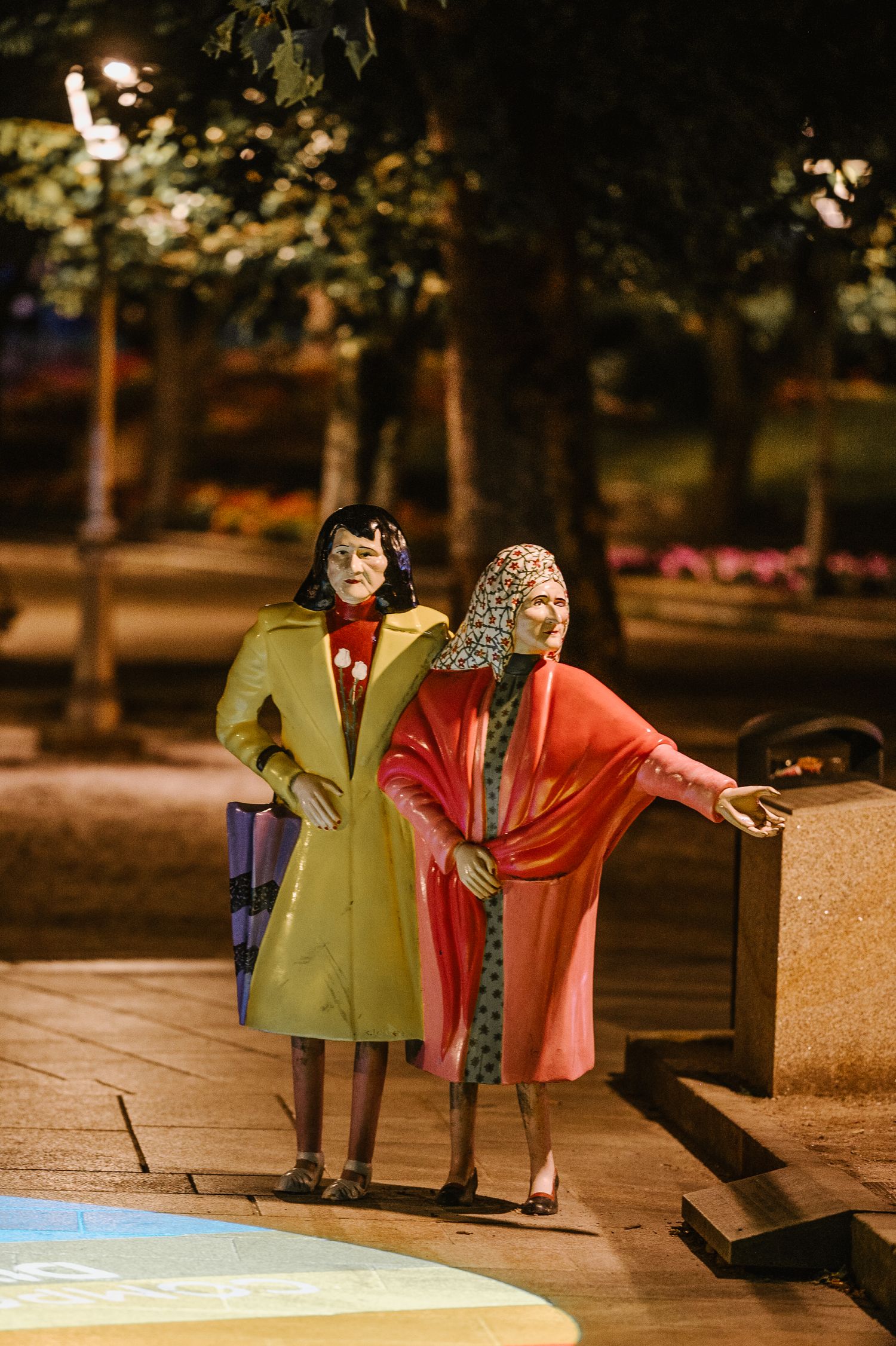 Two colorful sculptures of figures in bright jackets sit on a bench in a dimly lit park at dusk.