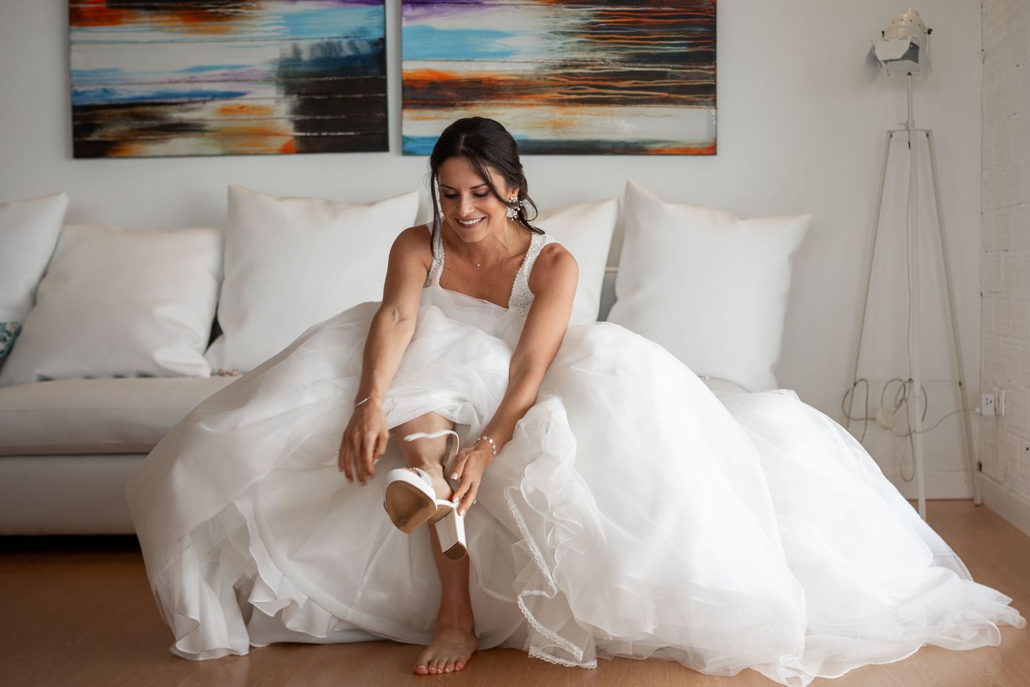 Bride in white wedding dress sitting on couch putting on shoes with colorful artwork in background.