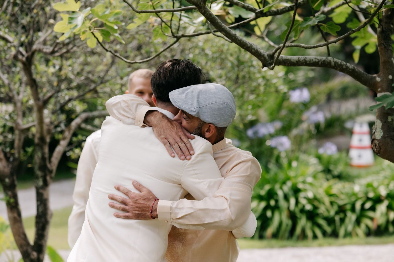 Two people share a warm embrace outdoors in a garden setting with trees and purple flowers in the background.