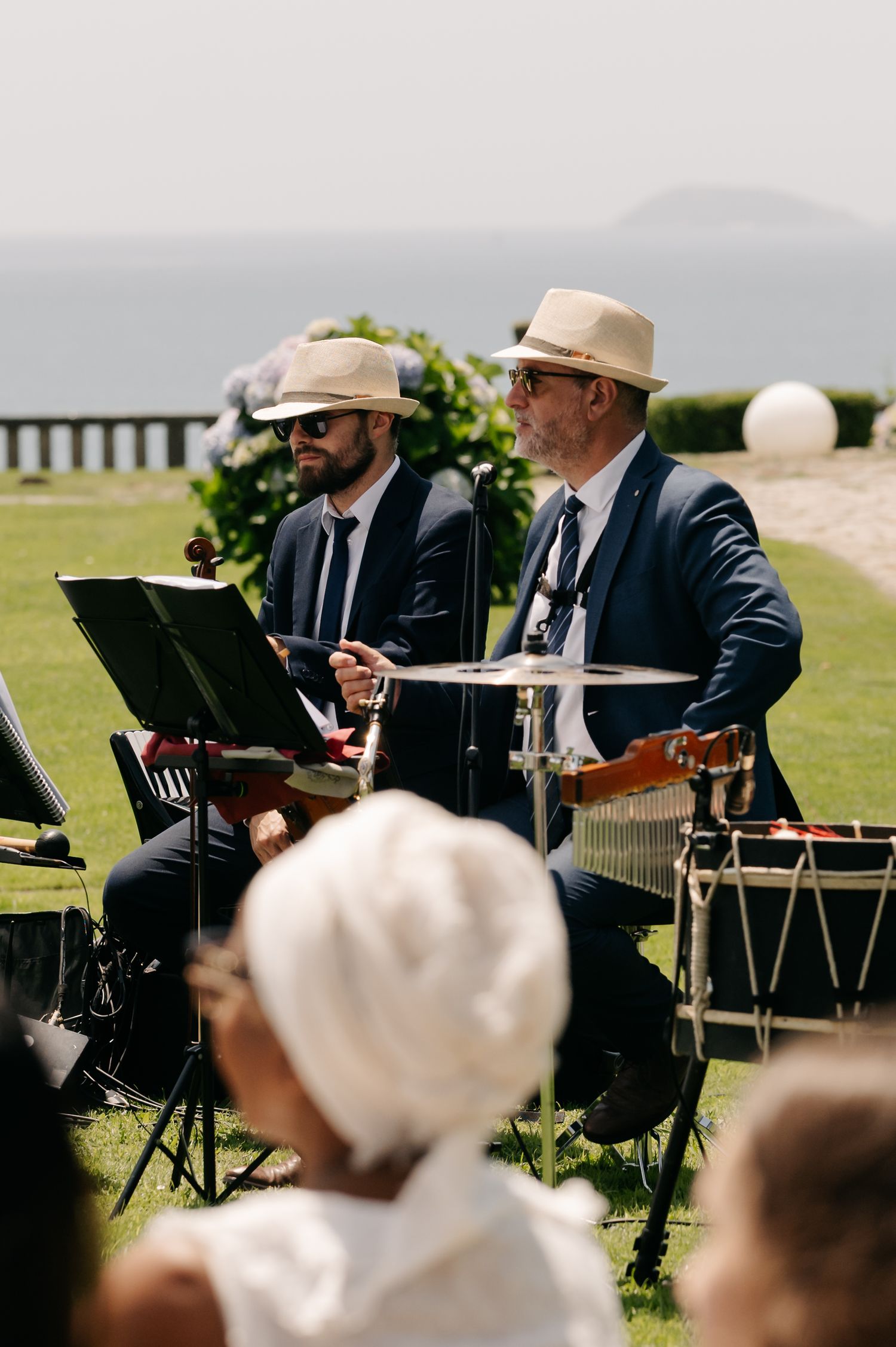 Musicians in suits and hats perform at an outdoor event with drums and instruments against a scenic backdrop.