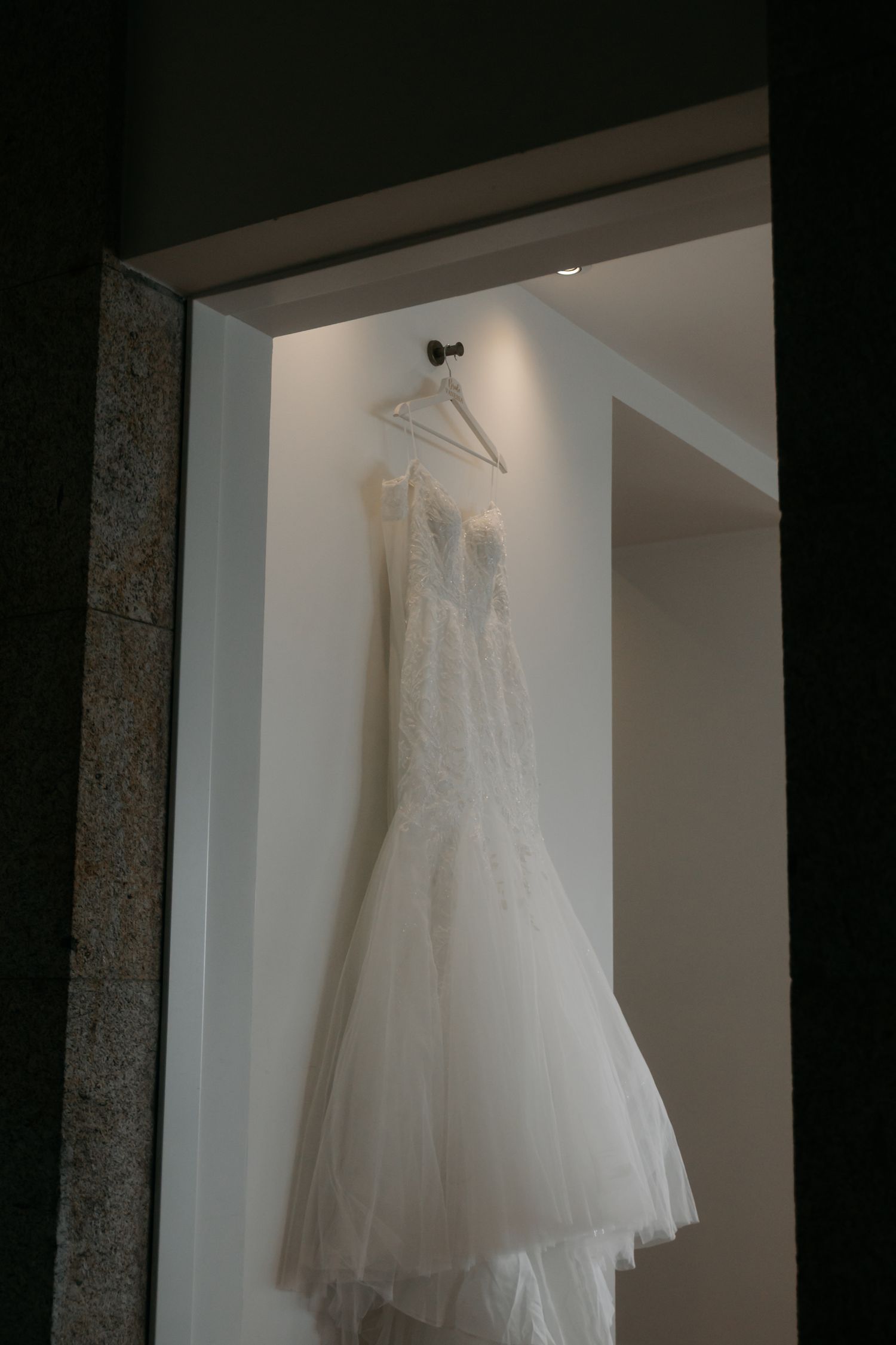 A white wedding dress with delicate lace detailing hangs elegantly in a window alcove against a dark background.