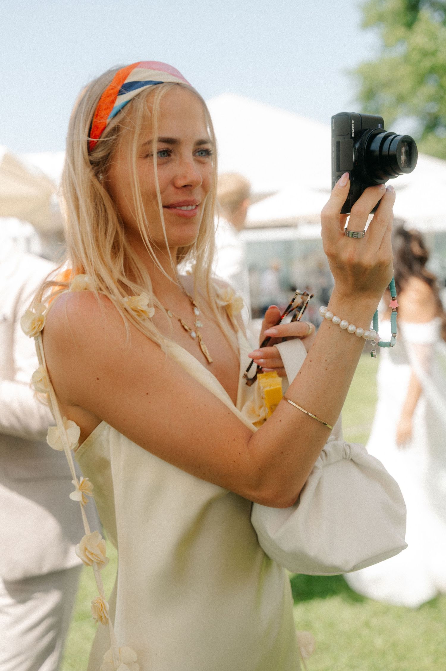 A photographer in a cream-colored dress takes photos outdoors with a camera during a sunny day.