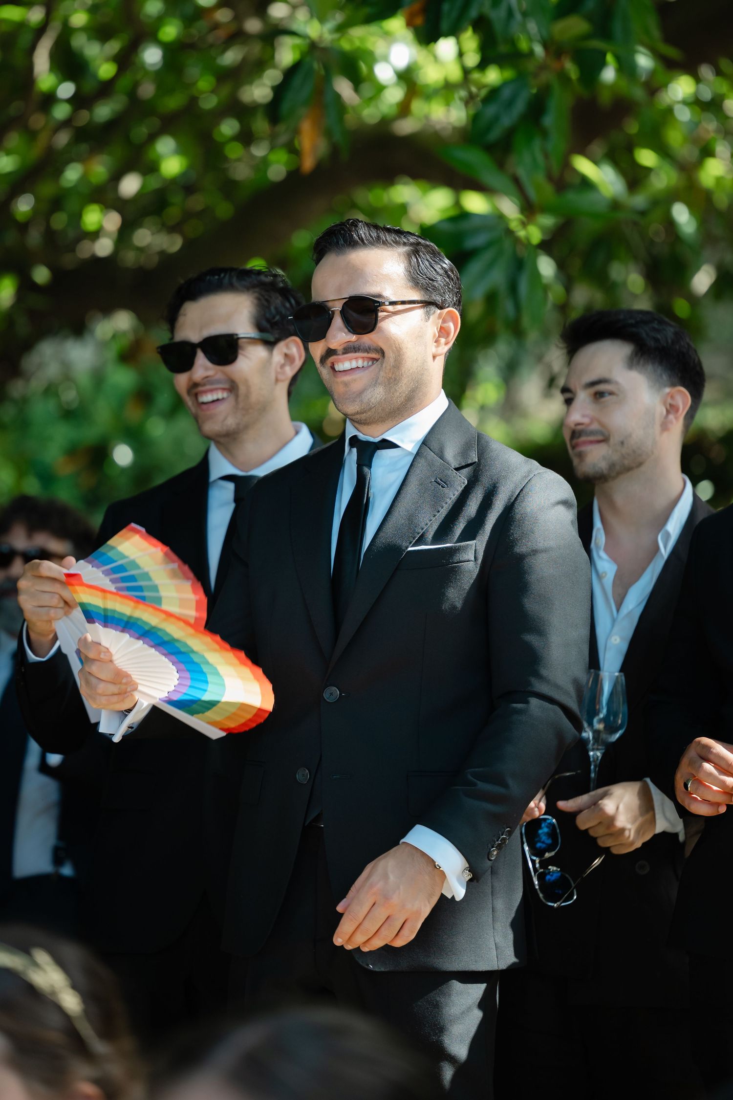 Three men in dark suits and sunglasses smile outdoors while one holds a rainbow flag at an event.