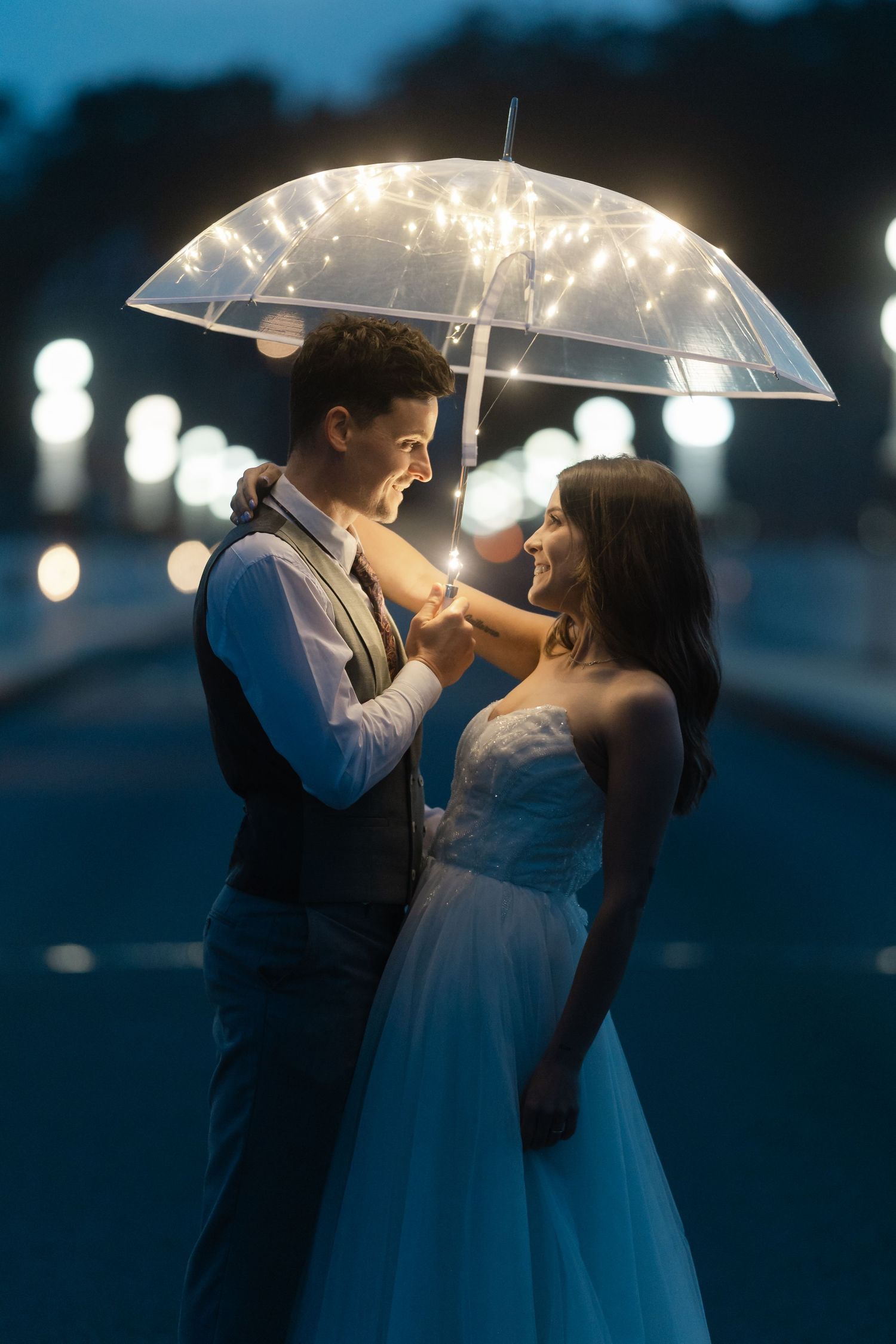 Couple sharing a romantic moment under a clear umbrella with fairy lights at dusk.