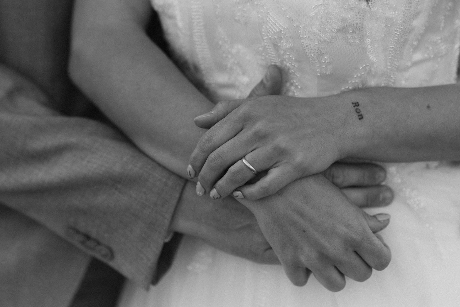 Black and white photo of newlyweds holding hands, showing wedding rings and a small tattoo, bride in white dress.