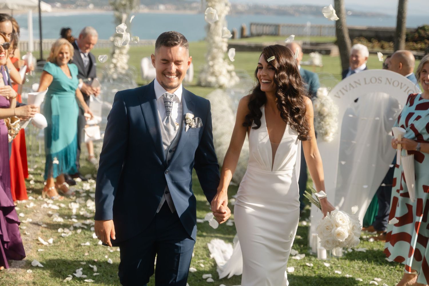 Wedding couple walks hand in hand through flower petals as guests celebrate at outdoor ceremony.