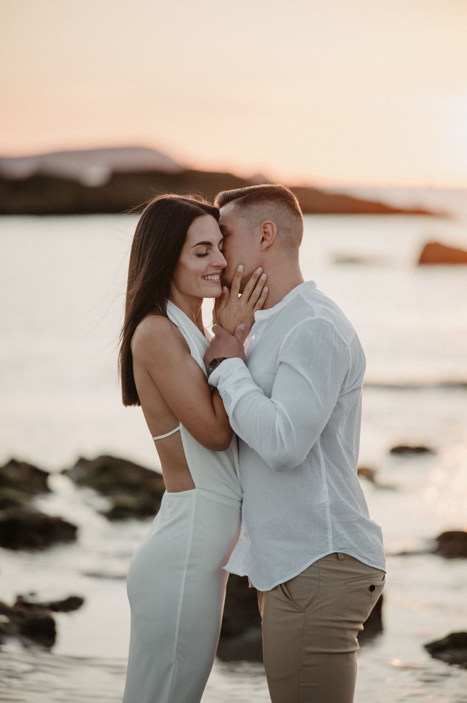Tender embrace between couple in white at sunset by rocky coastal shoreline.