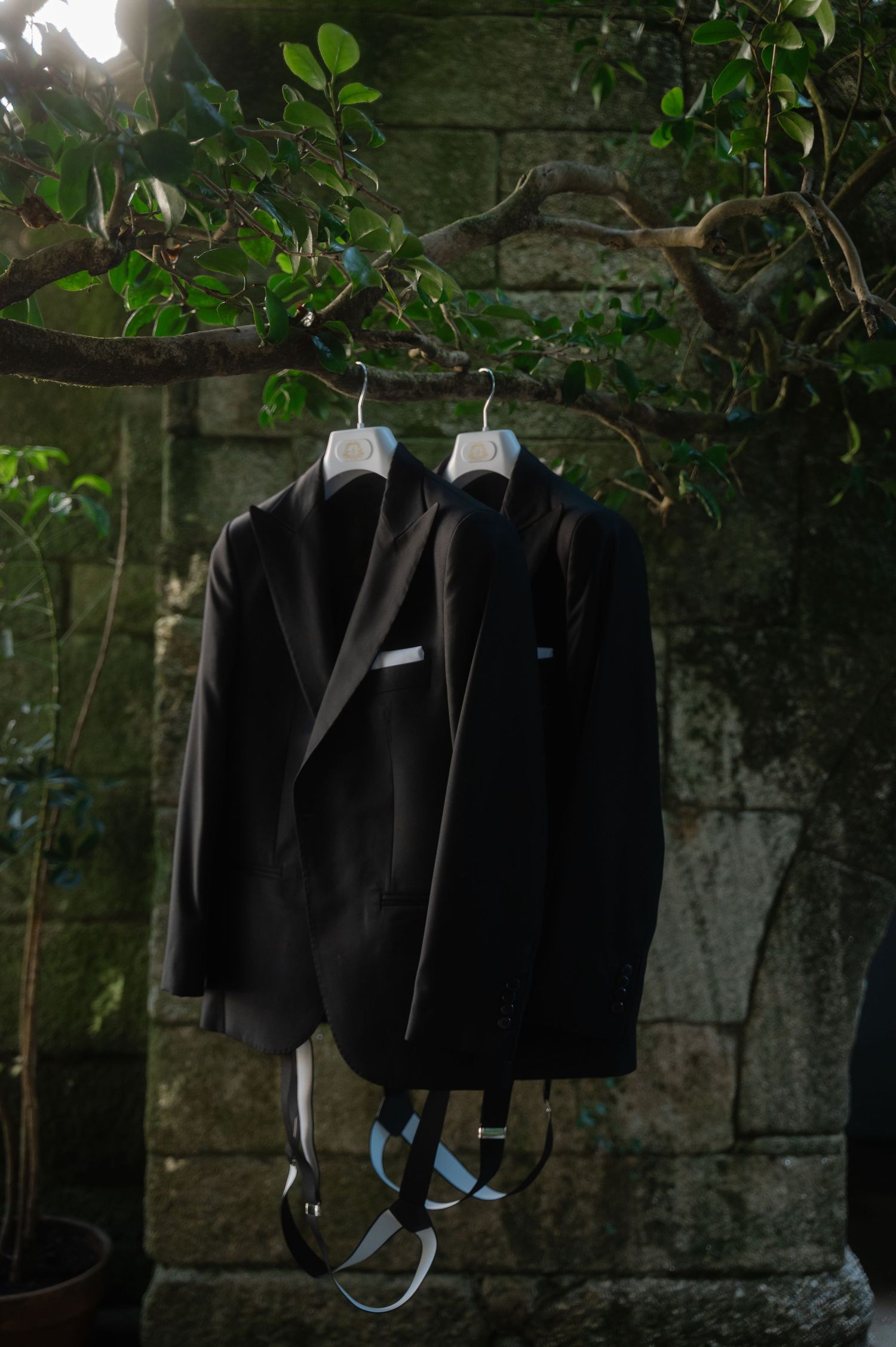 Black formal jackets hanging on hangers against a stone wall with green foliage in the background.