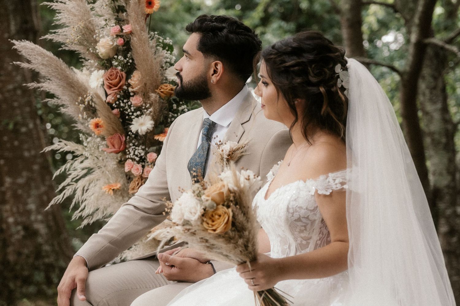 A couple sits together at their outdoor wedding ceremony surrounded by pampas grass and floral arrangements.