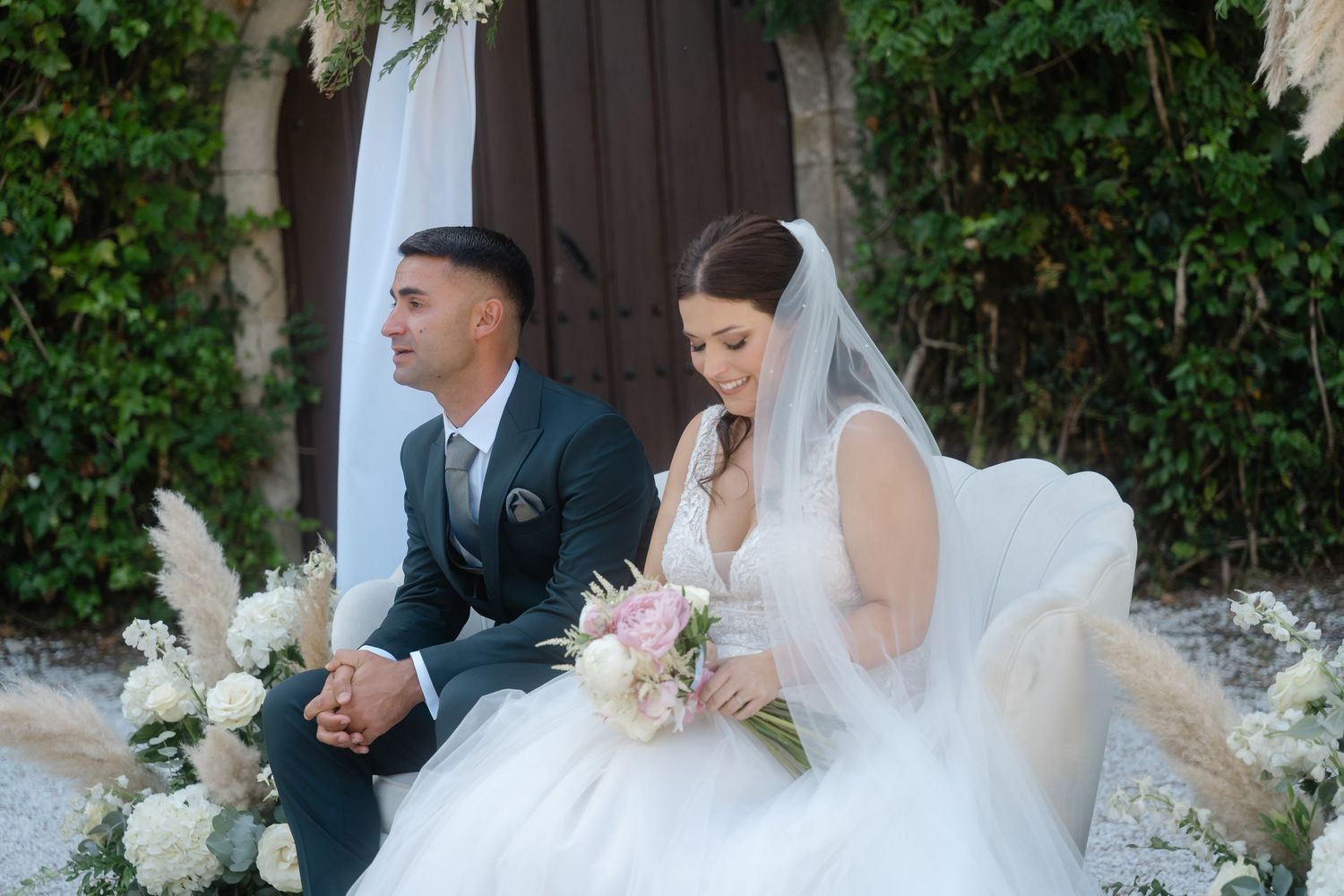 A couple sits together at their wedding ceremony with pampas grass and floral decor against a rustic backdrop.