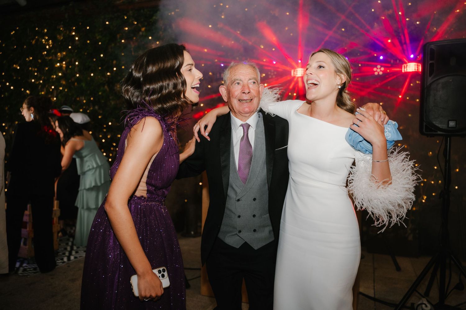 Wedding party dancing and celebrating on a dance floor illuminated by vibrant purple and red lights.