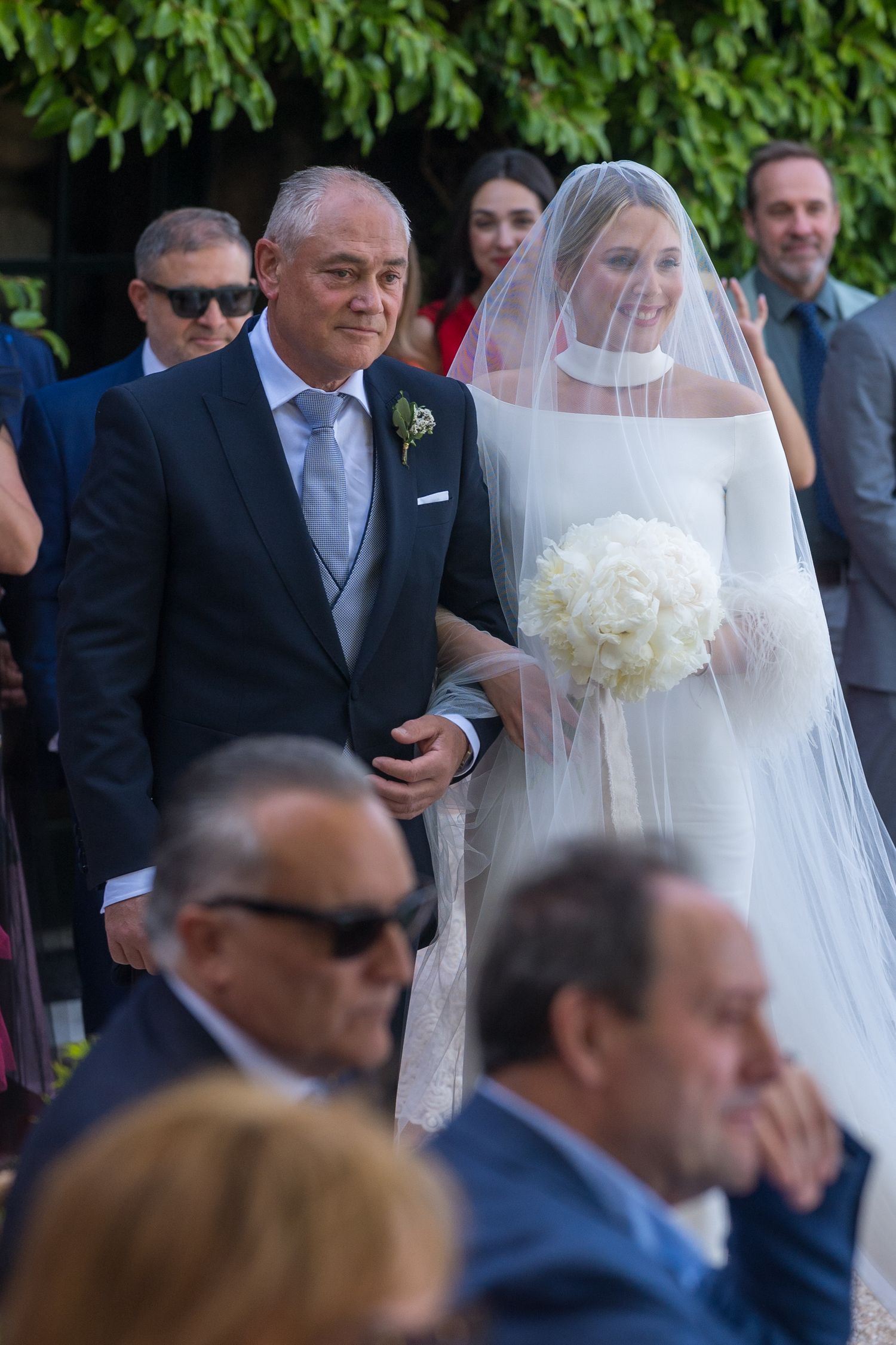 A wedding processional outdoors with guests watching as a bride in white veil carries a bouquet.