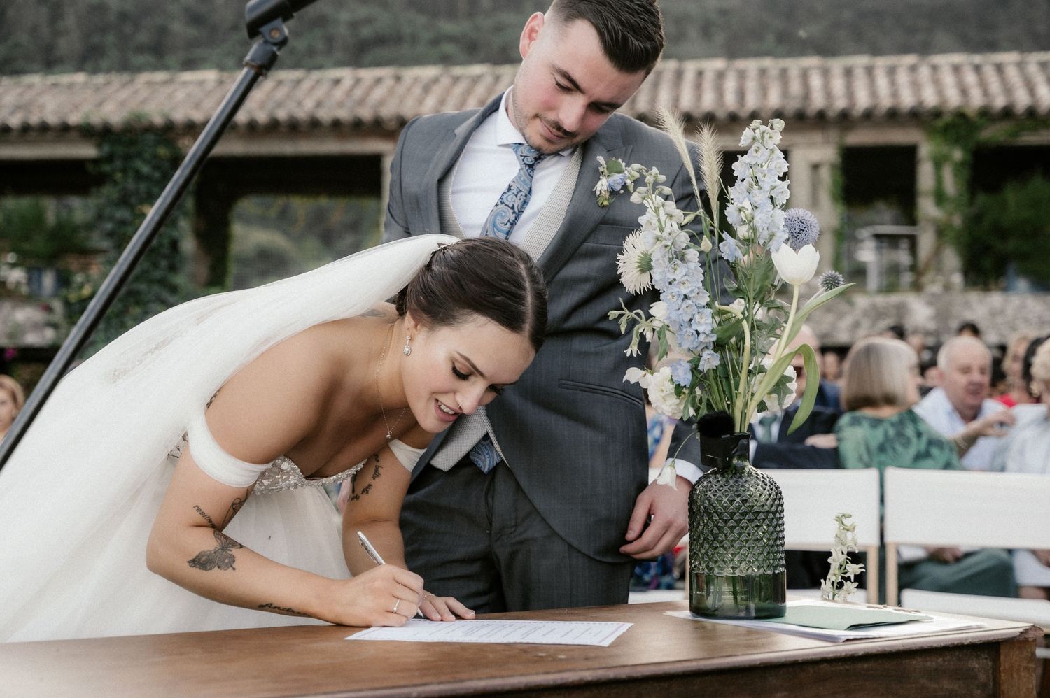 A bride in white signs wedding documents at a wooden table during an outdoor ceremony.