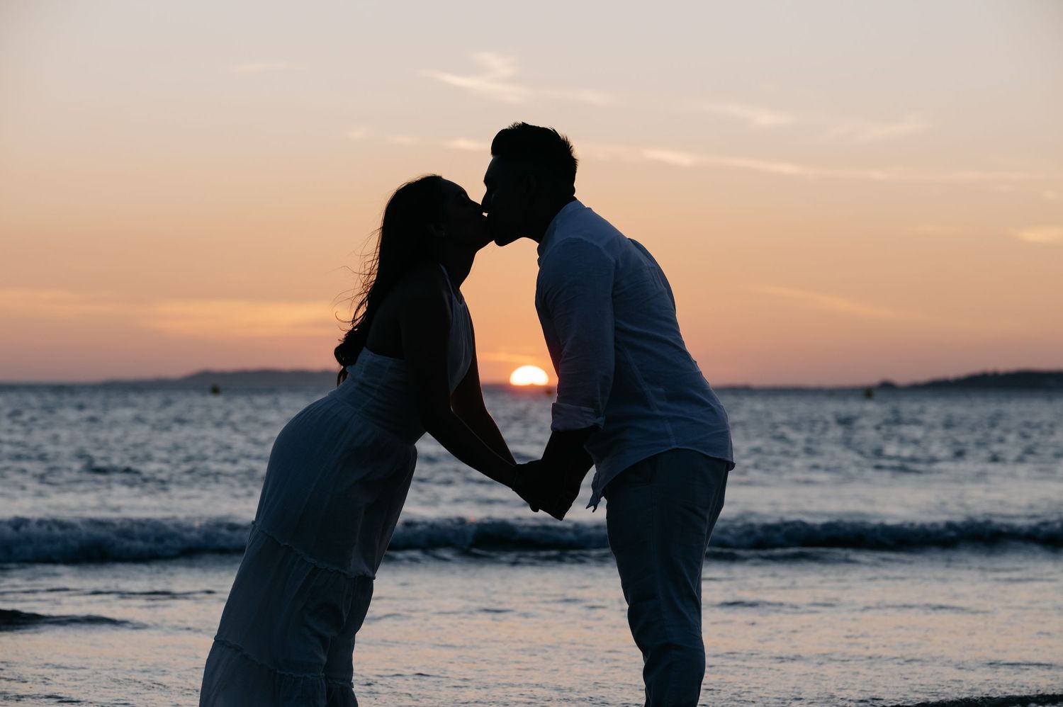 Silhouettes of a romantic couple share an intimate moment against a vibrant orange sunset at the beach.