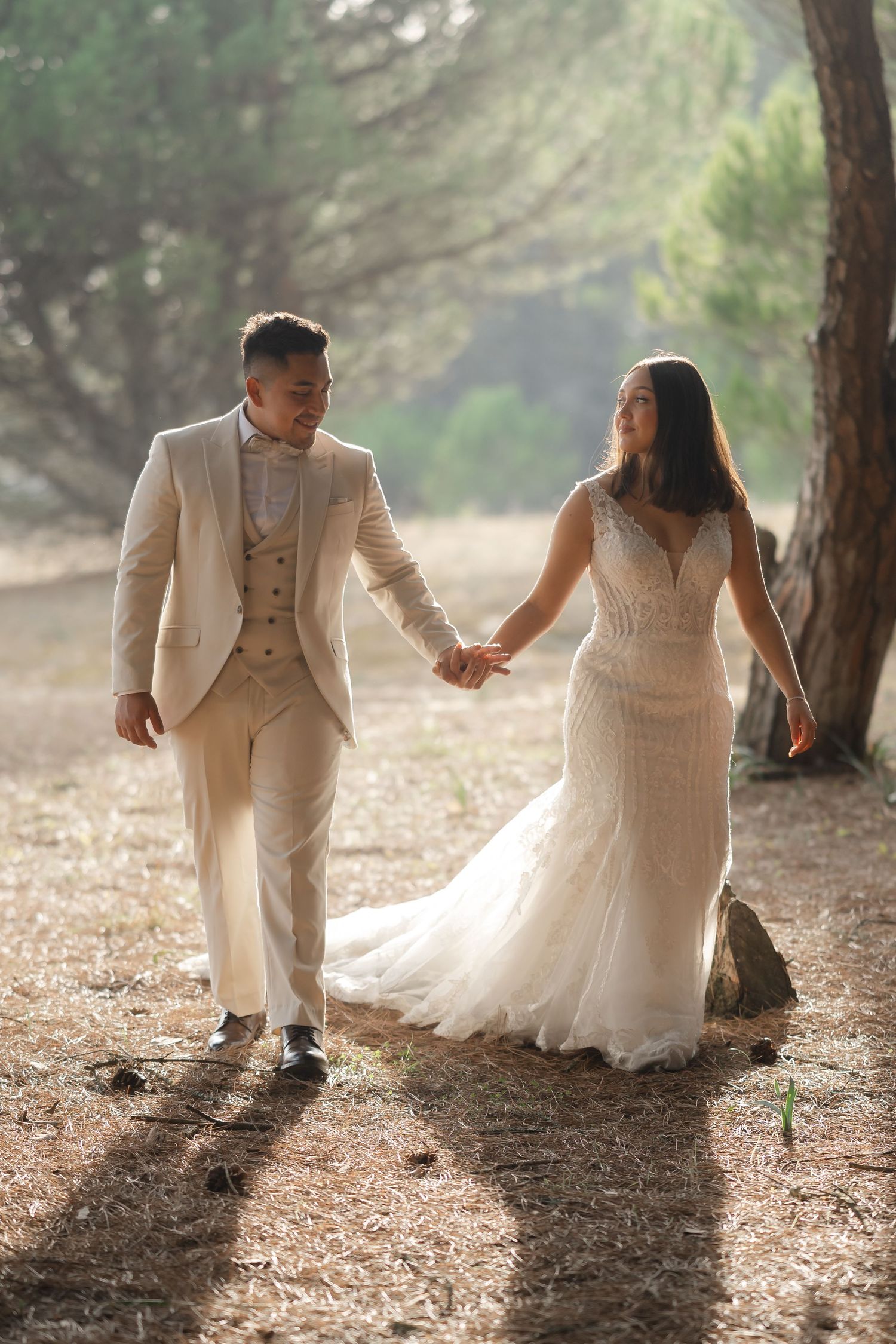 A wedding couple walks hand in hand through a sunlit forest path wearing cream and white formal attire.