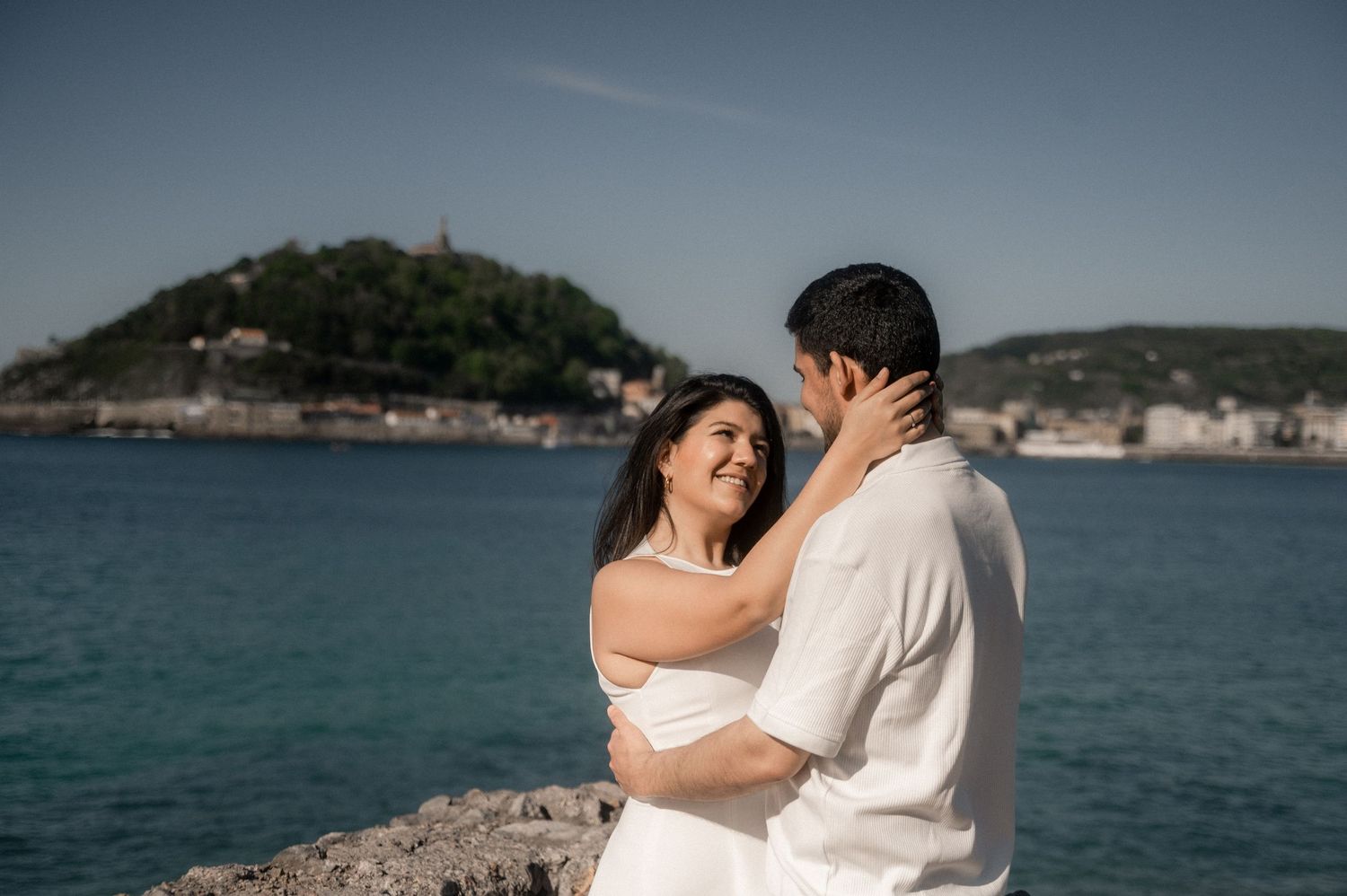 A couple embraces romantically on a beach with the ocean and coastline in the background during a sunny day.