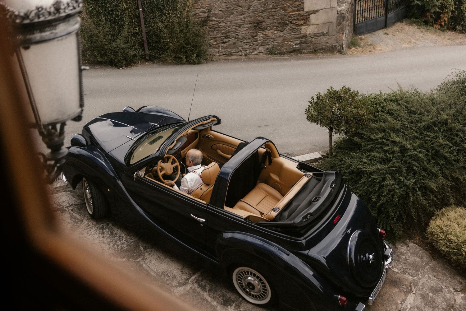 A classic black vintage convertible car parked on a stone driveway near lush greenery.