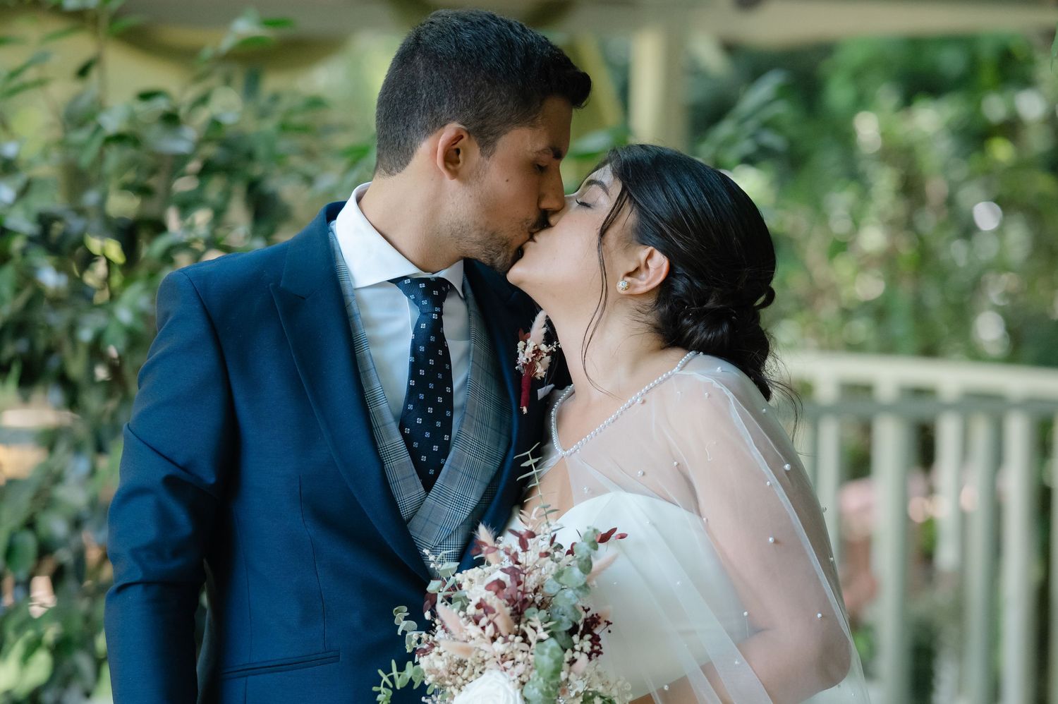 A romantic wedding couple shares tender moments in a garden setting with pink floral bouquets and a navy blue suit.