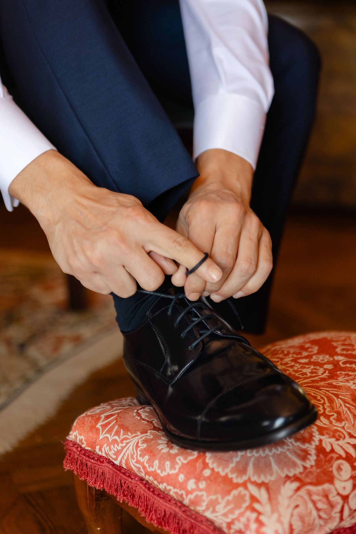 A person in a navy suit ties the laces of shiny black dress shoes resting on a red and white floral footstool.